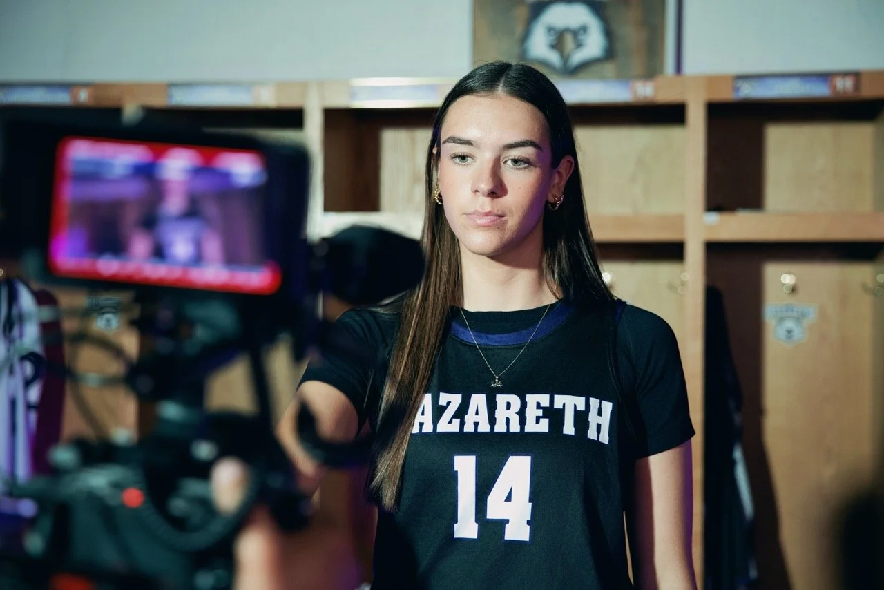 A young female athlete with long dark hair wearing a black basketball jersey with the name "AZARETH" and the number "14". She is in a locker room, being filmed or photographed, with wooden lockers in the background.