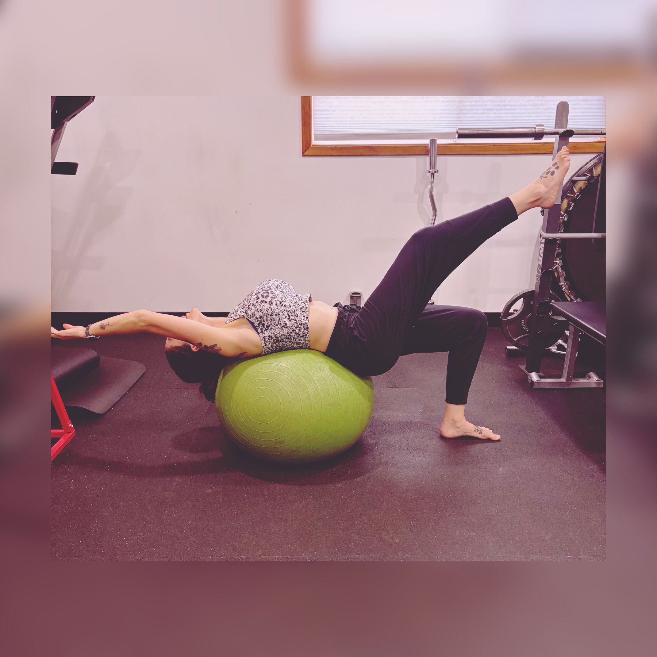 Cassie from CK Fitness, specializing in women's fitness, perimenopausal, pre and post natal and post menopausal fitness, performing a bridge on a green stability ball in a gym, with her arms extended overhead and one leg raised against a wall.