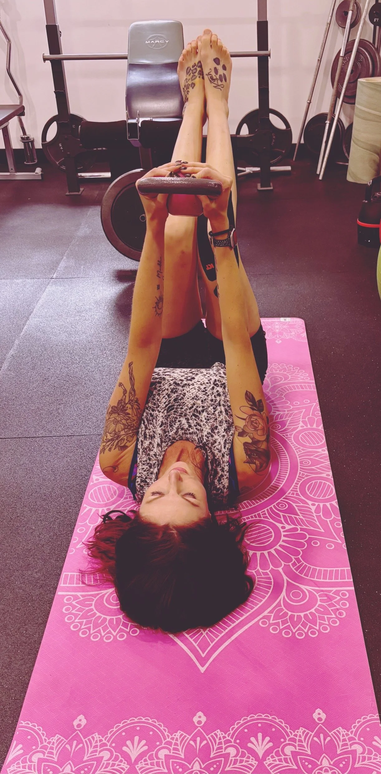Cassie from CK Fitness, specializing in women's fitness, perimenopausal, pre and post natal and post menopausal fitness, lying on a pink yoga mat lifting legs with dumbbell in gym.