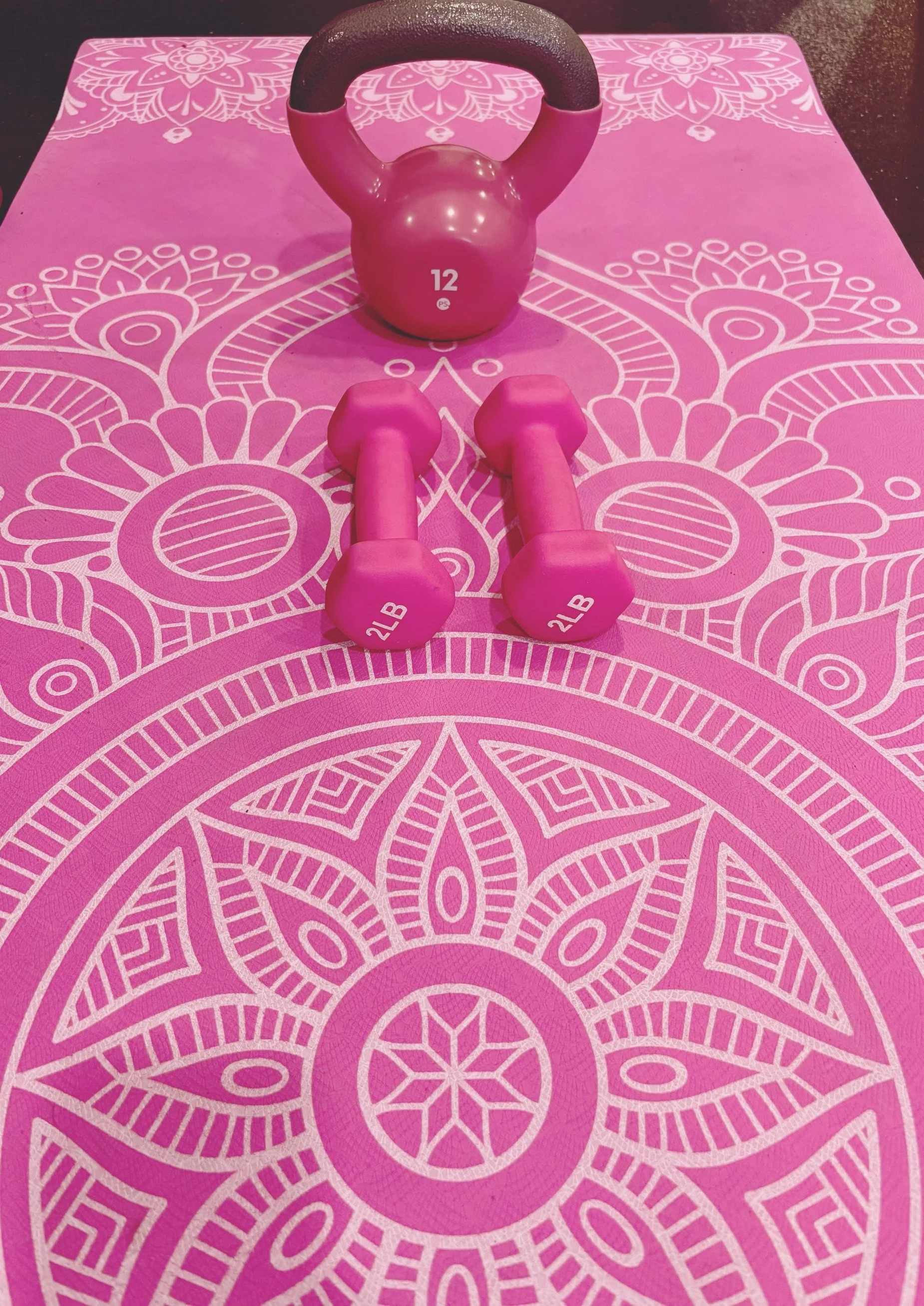 Pink yoga mat with white mandala pattern, pink kettlebell, and pink dumbbells on it.