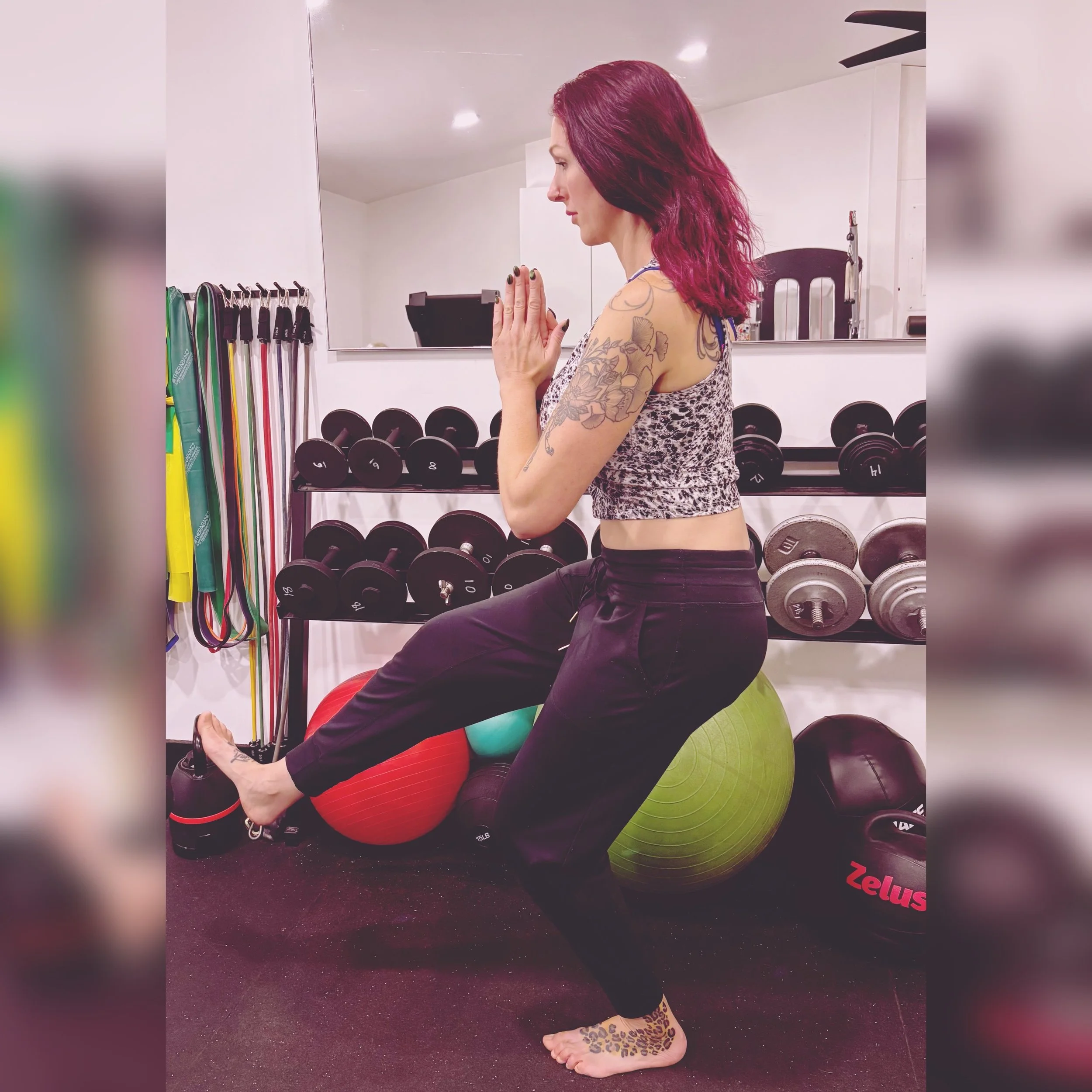 Cassie from CK Fitness, specializing in women's fitness, perimenopausal, pre and post natal and post menopausal fitness, practicing yoga on a stability ball in a gym, with dumbbells, resistance bands, and other exercise equipment in the background.