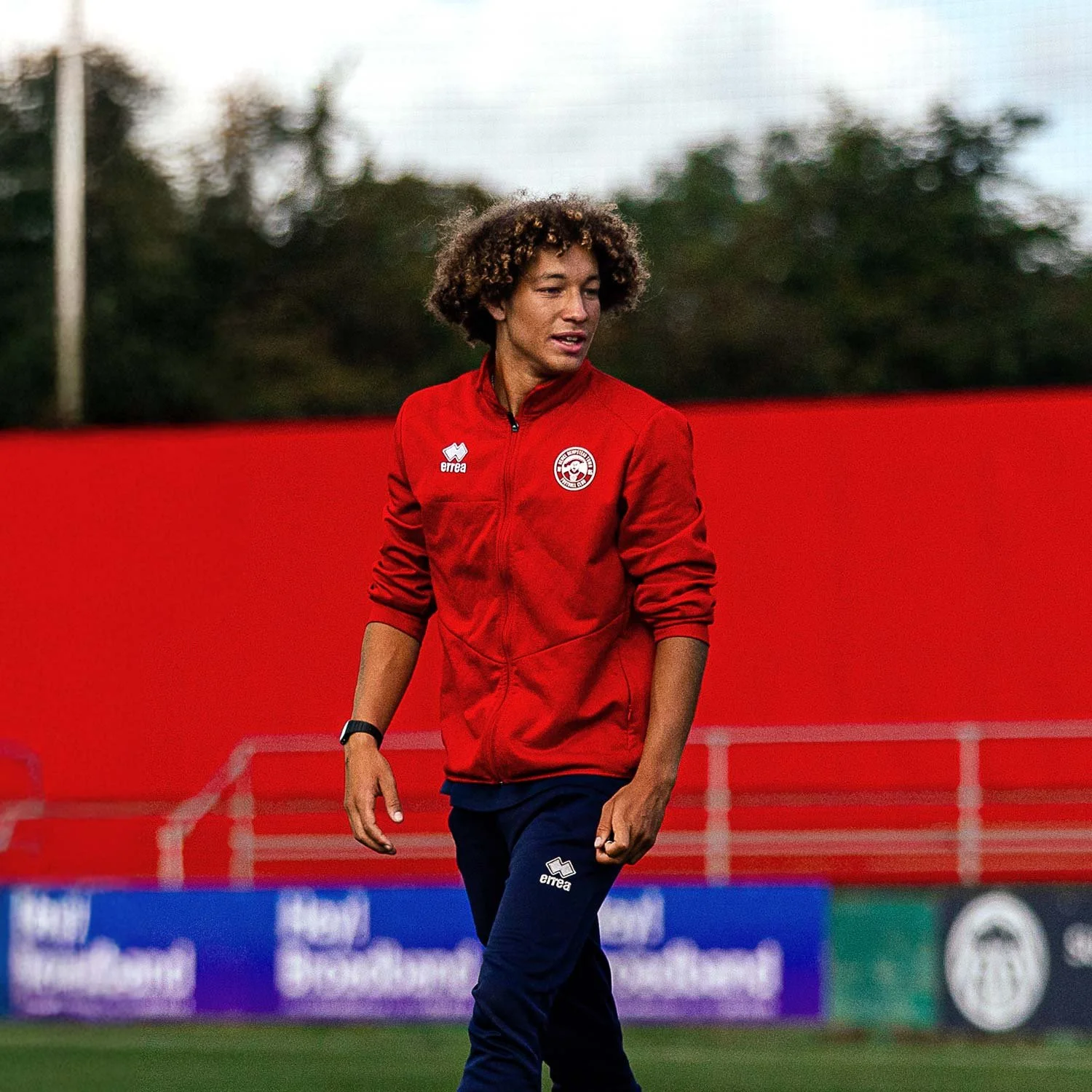 A woman with curly hair wearing a red sports jacket and navy athletic pants walking on a soccer field.