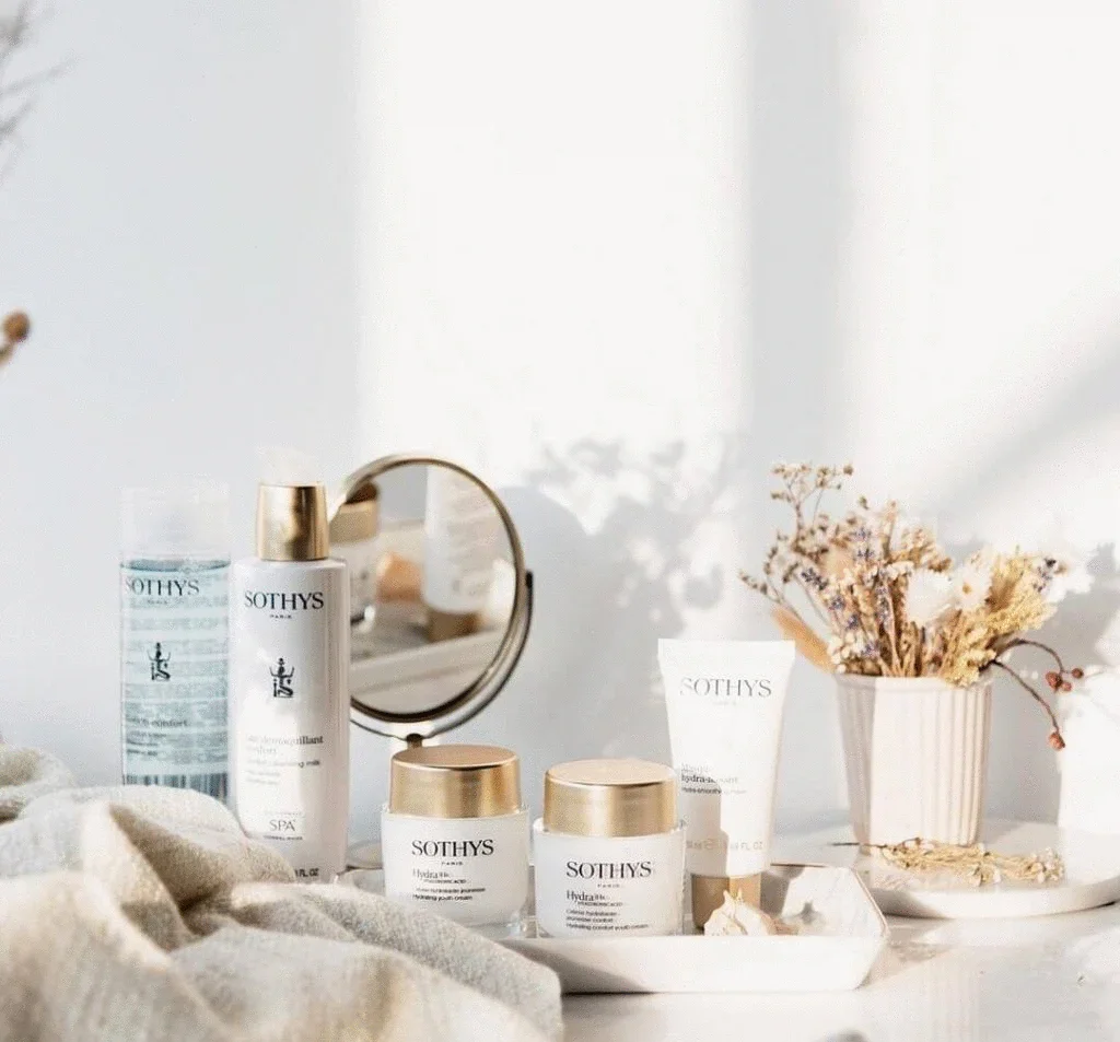 Produits de soin de la peau SOTHYS disposés sur une table blanche avec un miroir, un vase avec des fleurs séchées, et un tissu beige, dans une pièce lumineuse.