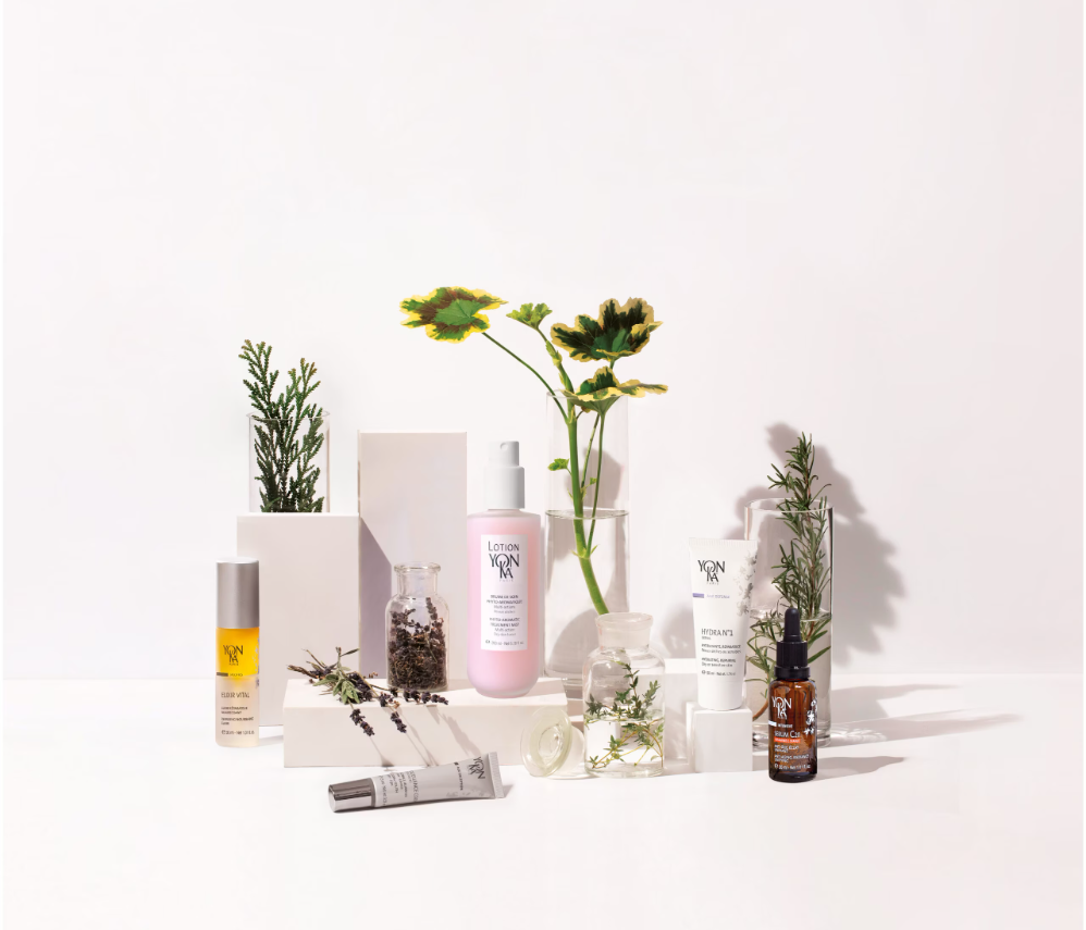 Produits de soins de la peau YON-KA et des huiles essentielles avec des plantes vertes sur un fond blanc.