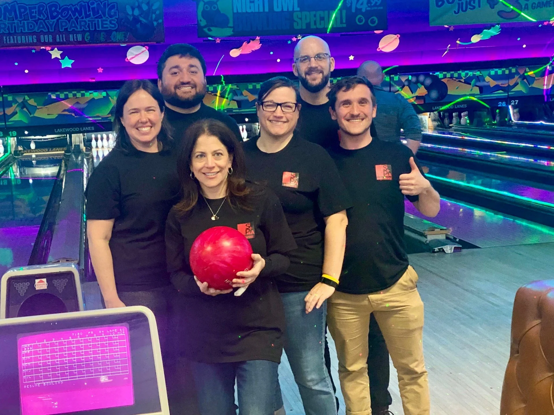 Group of six people at a bowling alley, smiling, one woman holding a red bowling ball, colorful neon lights, bowling lanes in the background.
