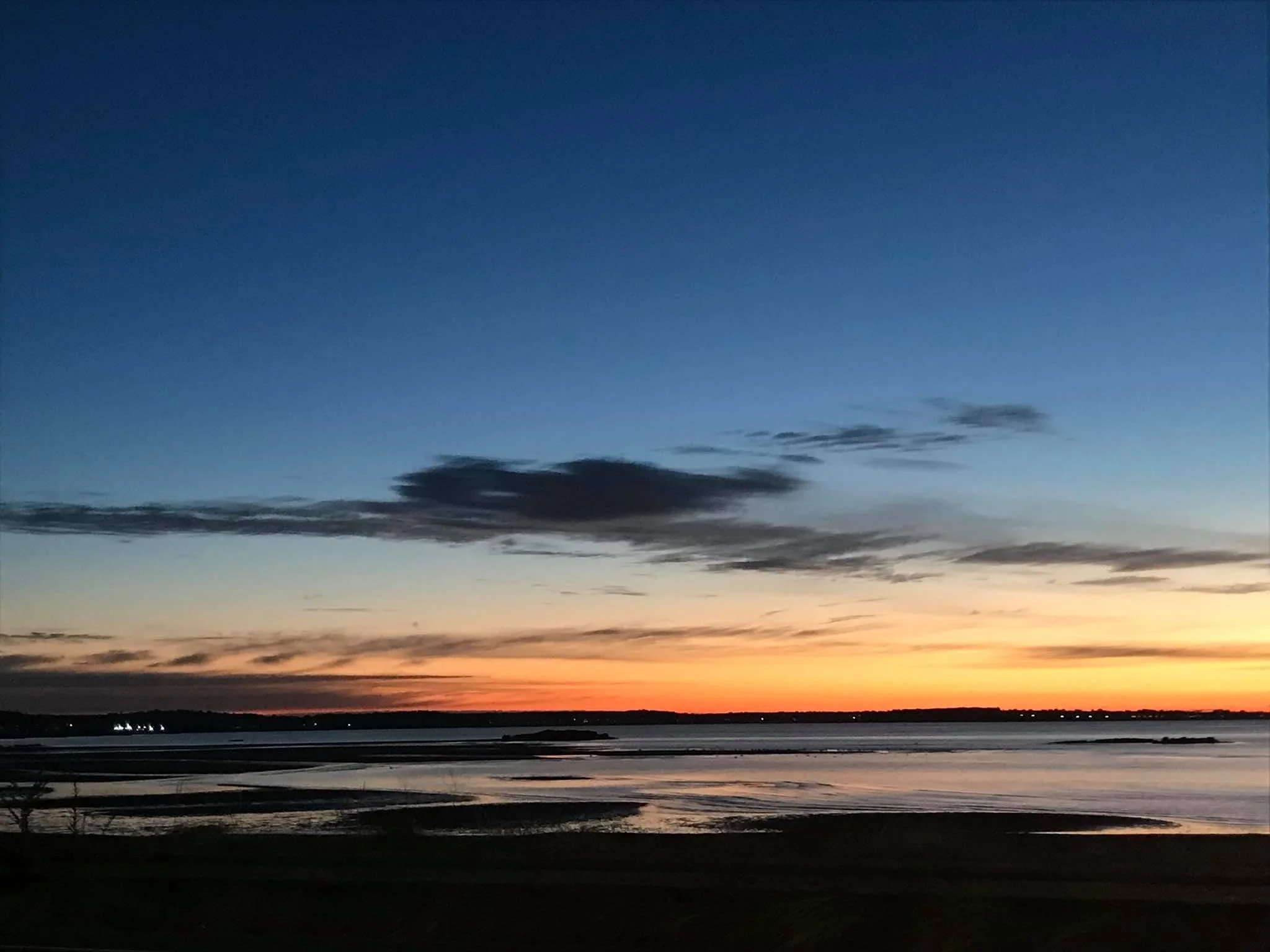 Sunset over a body of water with partly cloudy sky and distant lights on the horizon.