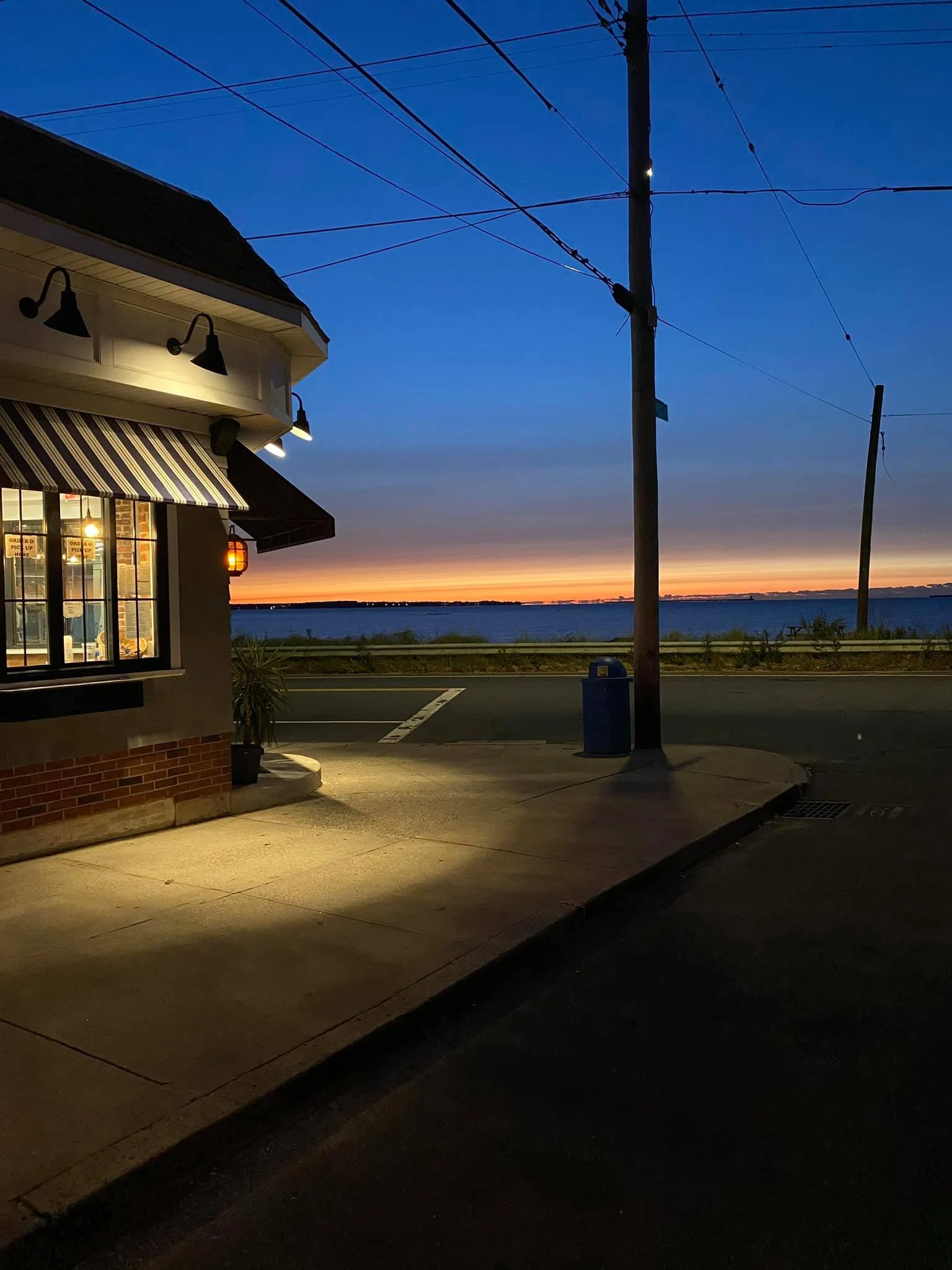 A view of a cafe building with outdoor lighting at sunset, overlooking a body of water, with a street, utility poles, and power lines in the foreground.