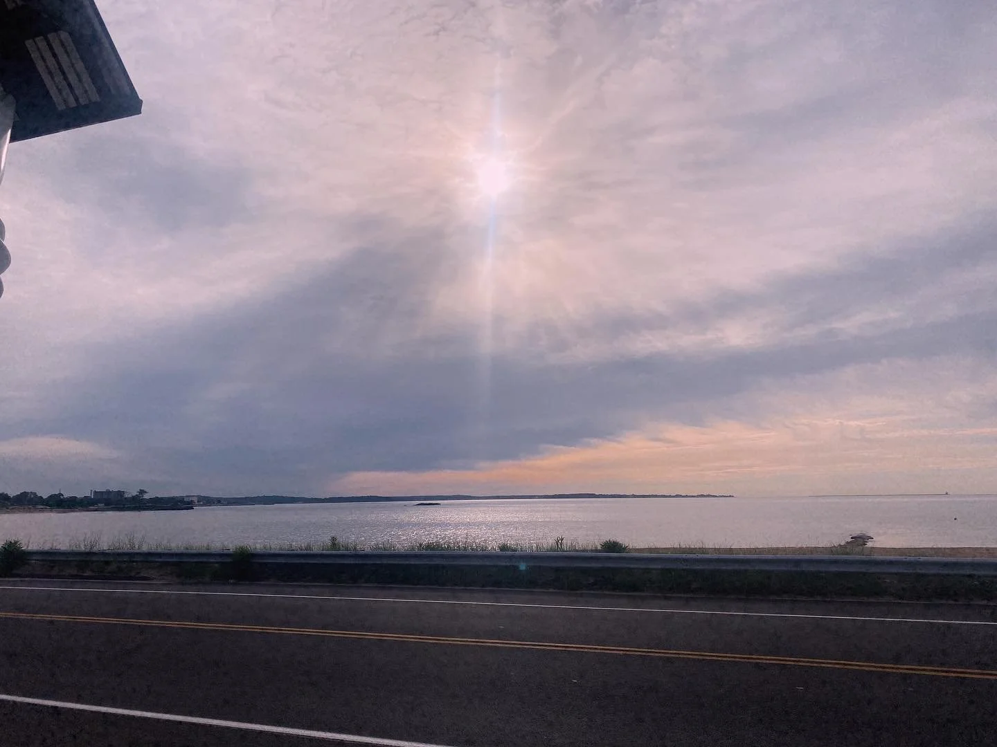 Scenic view of a water body under a sky with sun and clouds, seen from a roadside with a guardrail.