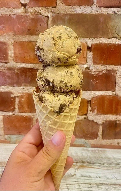 Hand holding an ice cream cone with three scoops of pistachio-flavored ice cream against a brick wall background.
