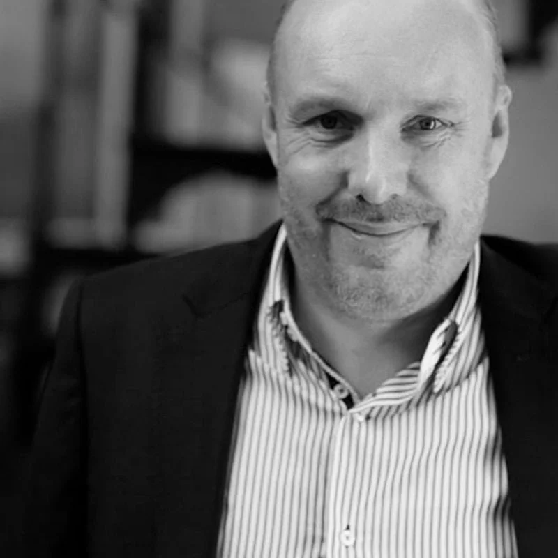 Black and white photo of a smiling man in a suit jacket and striped shirt, with a blurred background of bookshelves.