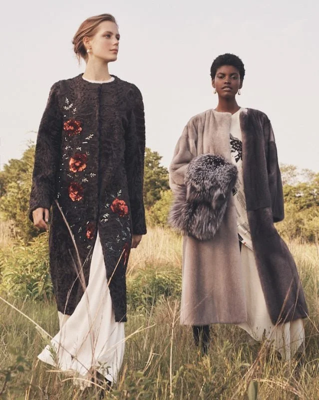 Two women standing in a grassy field, wearing stylish long coats with one holding a fur accessory, under a cloudy sky.