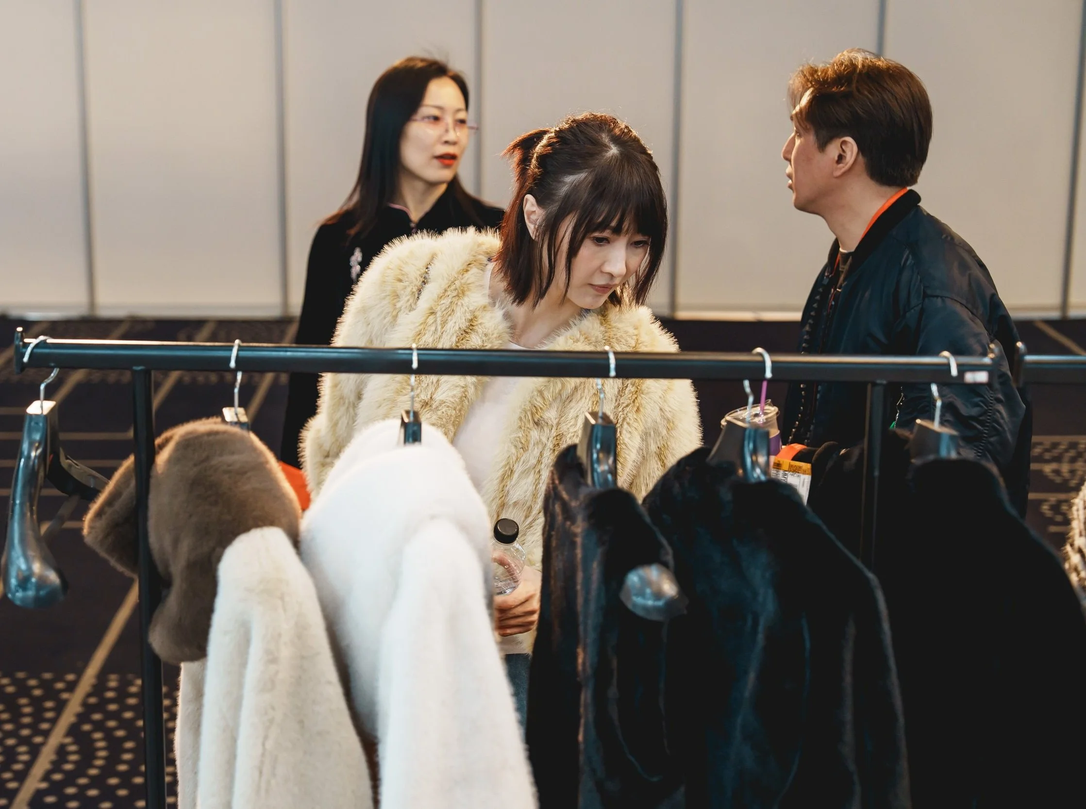 Three people shopping at a clothing booth with fur coats on hangers, in an indoor space.