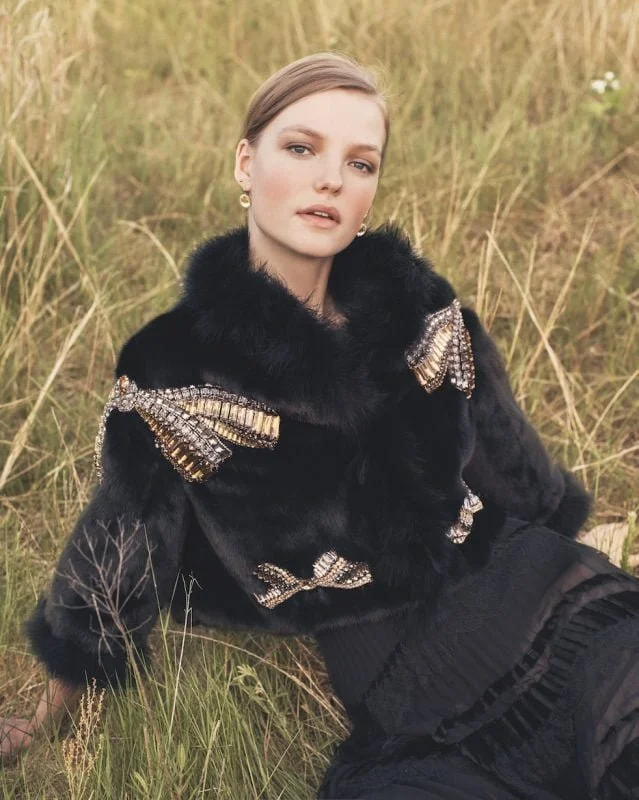 A woman wearing a black fur coat with embellished gold and silver details, sitting in a grassy field.