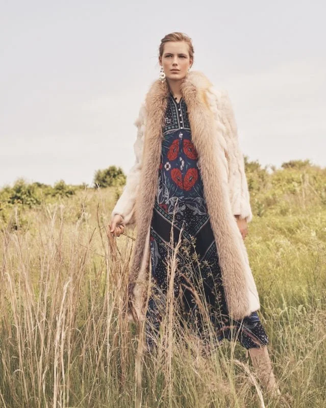 A woman with blonde hair standing in a grassy field wearing a long, cream-colored fur coat over a blue dress with red floral embroidery.