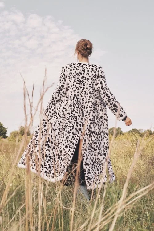 Woman wearing a long leopard print coat standing in a grassy field with a partly cloudy sky in the background.