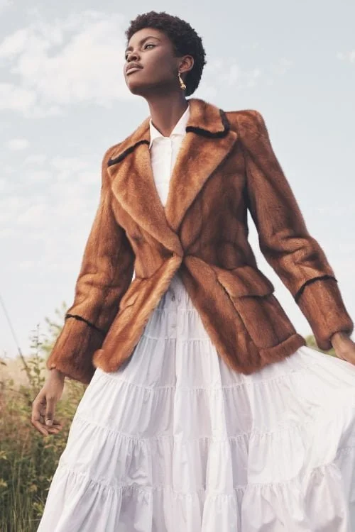 A woman wearing a brown fur coat and a white tiered dress stands outdoors against a cloudy sky.