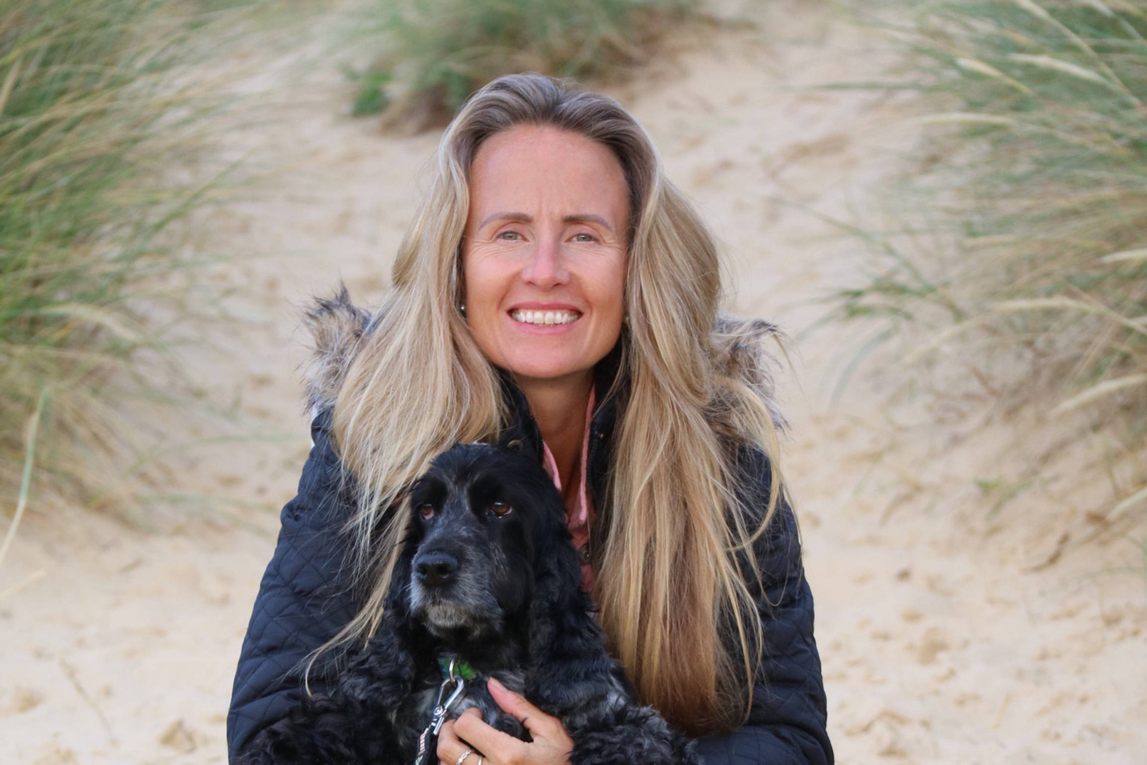 A smiling woman with long blonde hair holding a black dog on a sandy path surrounded by beach grass.
