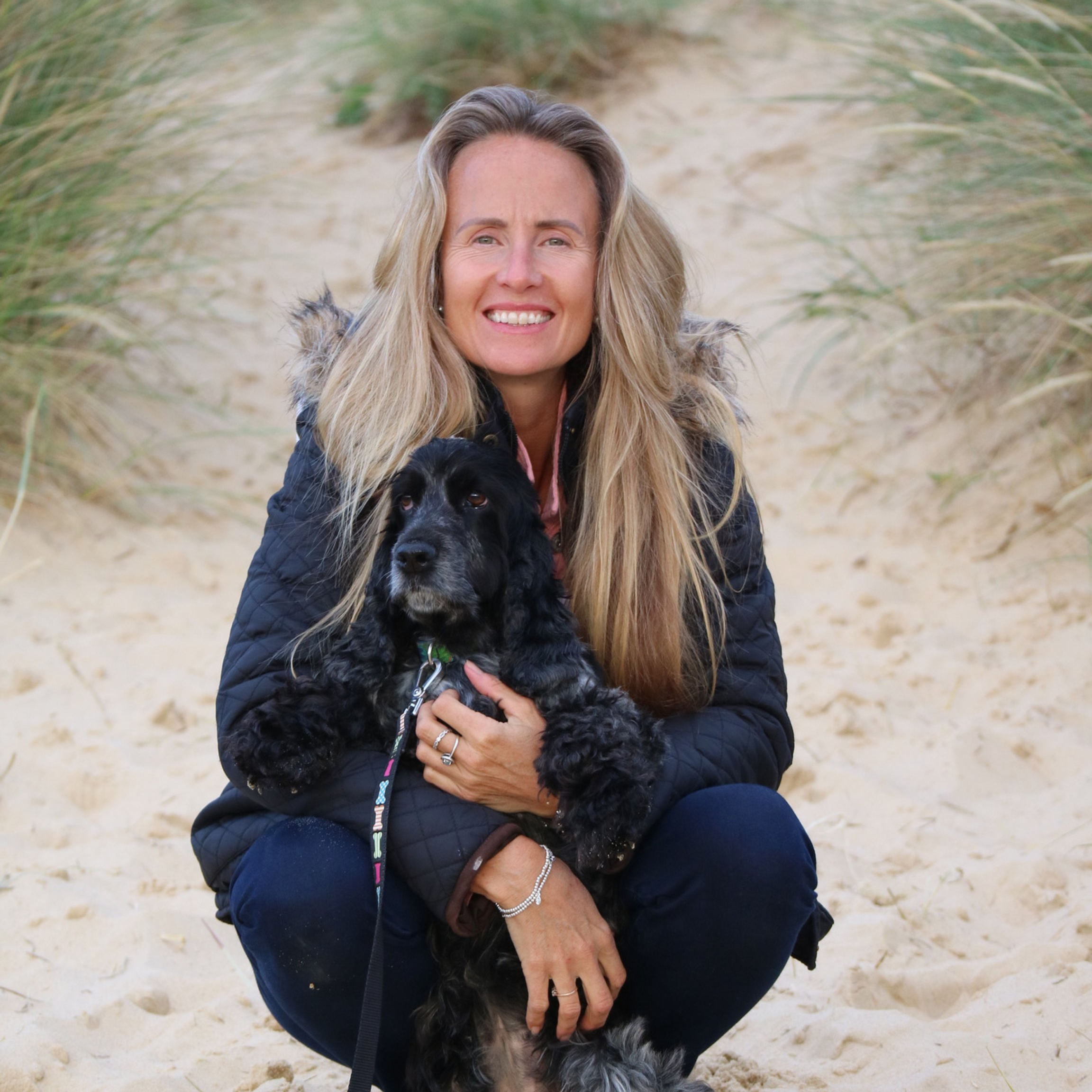 Suzi Hancock with long blonde hair smiling on a sandy beach grass path, sitting with a black and gray Cocker Spaniel dog.