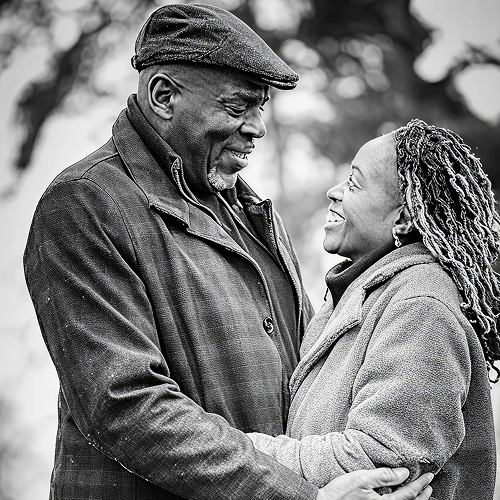 A man and a woman standing outdoors, close together, smiling at the camera with a blurred background of trees.