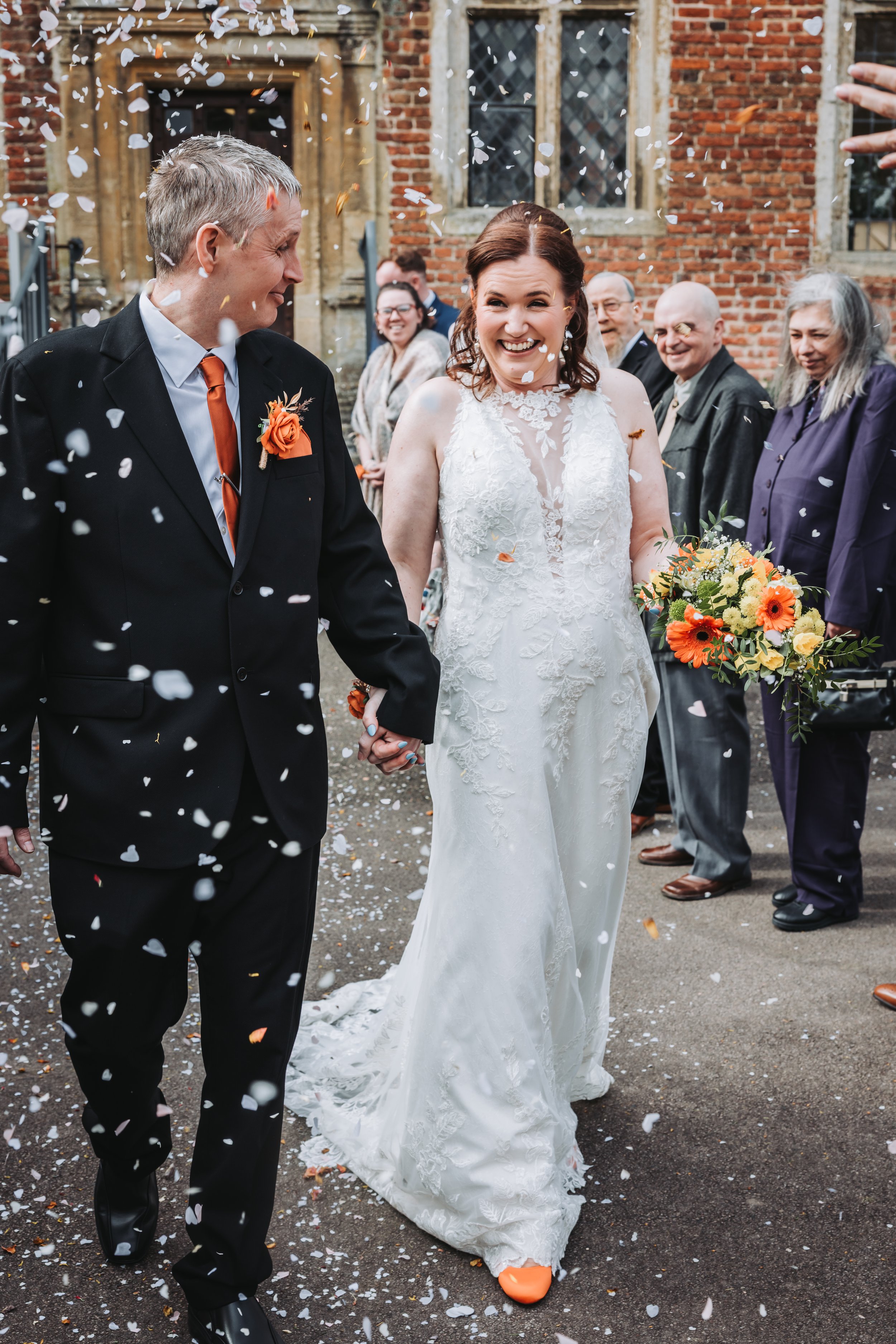 A couple on their wedding day, holding hands outdoors, with the bride in a white wedding dress and the groom in a light gray suit, smiling and standing in front of trees.