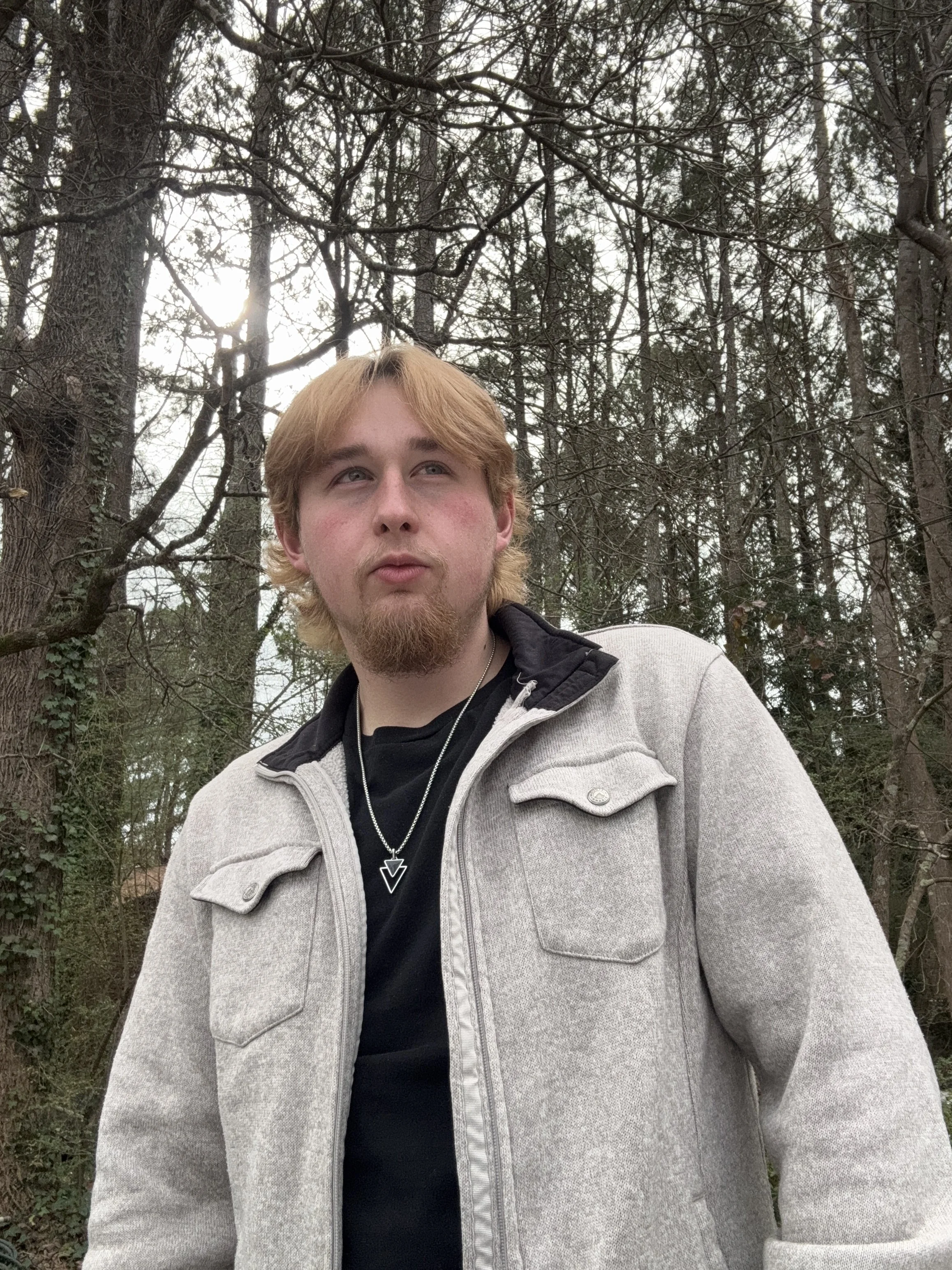 A young man with blonde hair stands near a lake on a wooden dock under a partly cloudy sky, holding sunglasses with one hand and wearing a gray T-shirt with a logo, a cross necklace, and a wristband.