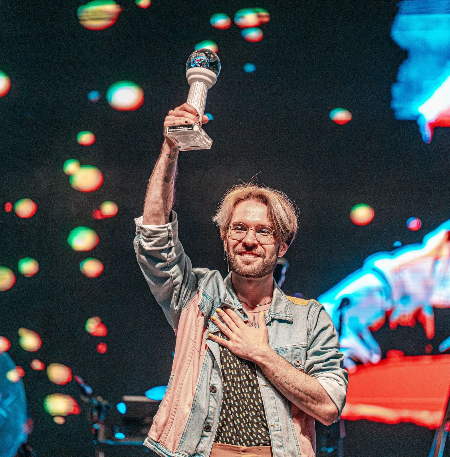 Homem sorrindo segurando um troféu com uma mão no peito, em um palco com luzes coloridas ao fundo.