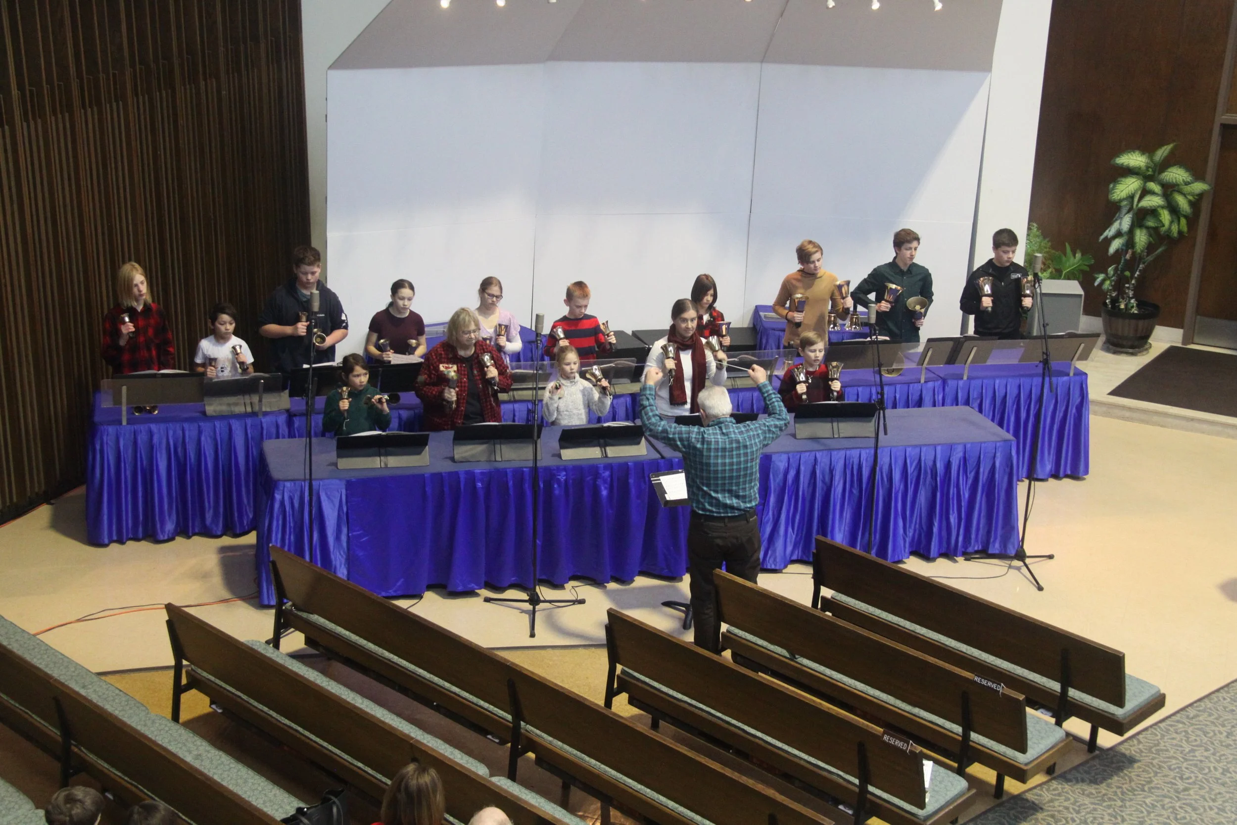 Children and teenagers performing a choir with a conductor in front, standing behind music stands on a stage with blue drapes, in a concert hall.