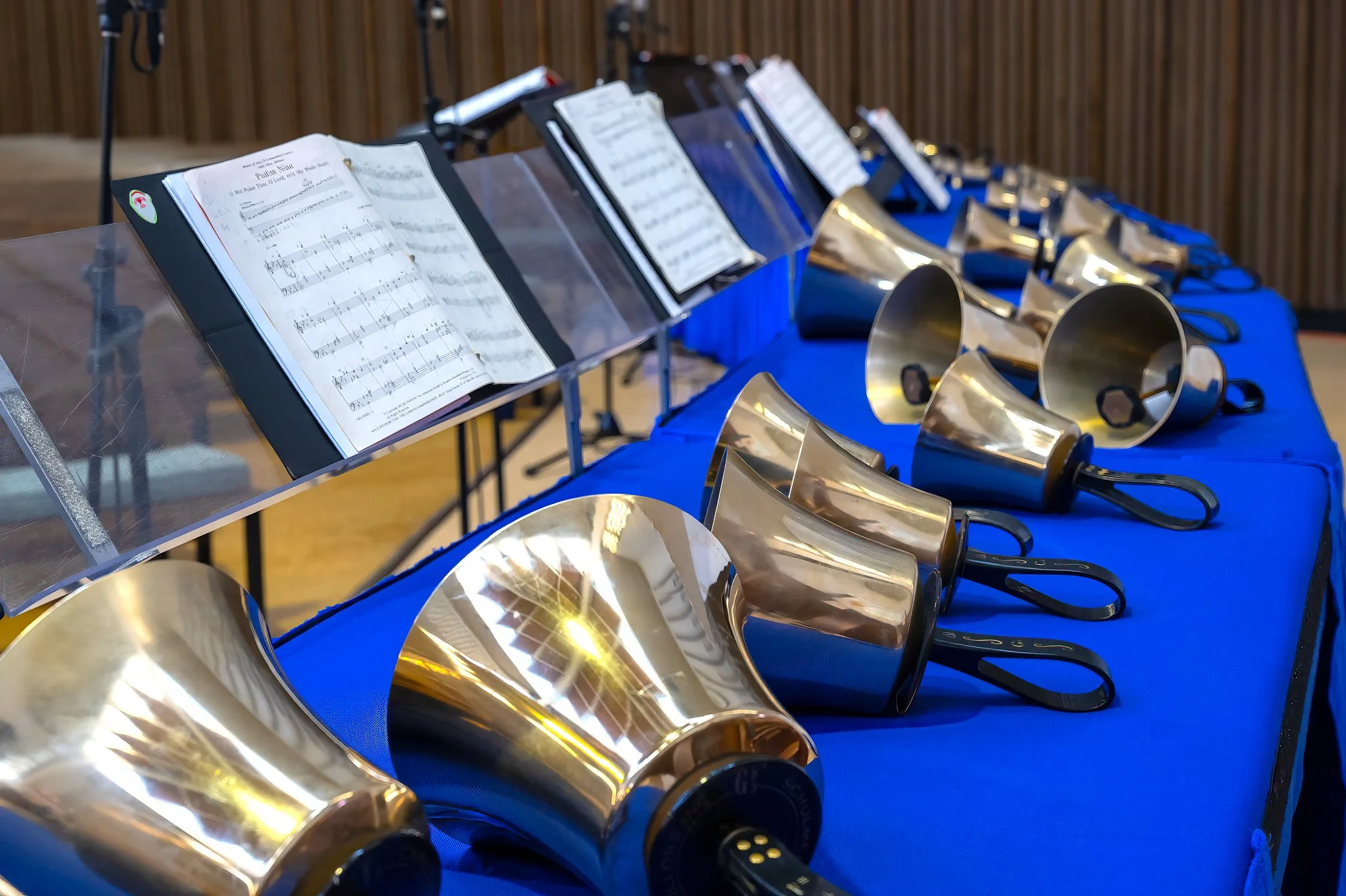 A row of brass handbells with black handles on a blue tablecloth, with sheet music stands holding open sheets music behind the table.