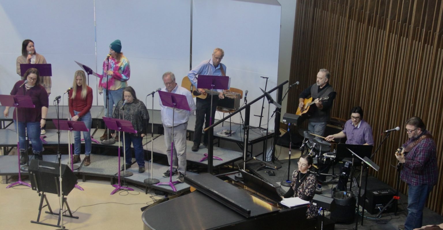 A group of people performing music and singing, with some playing guitars, a keyboard, drums, and others holding microphones and sheet music in a room with a wooden wall.