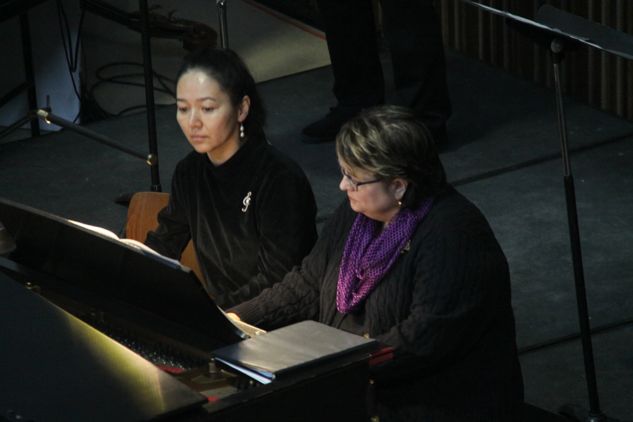 Two women are sitting at a black grand piano on a stage, playing music. One woman has dark hair tied back, wearing a black top, and the other has short hair with glasses and a purple scarf. There are music sheets on the piano.