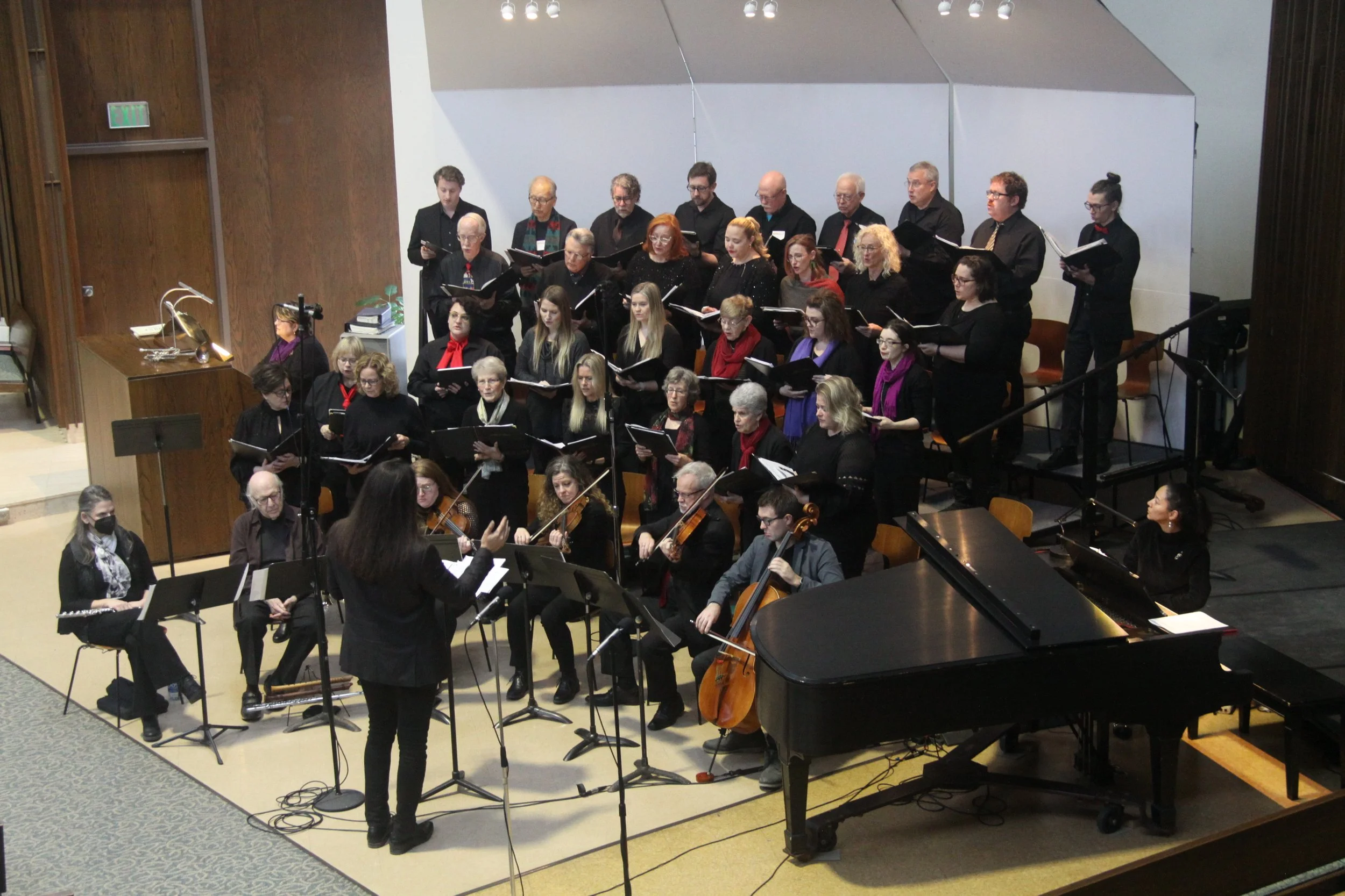 A choir and orchestra performing on stage, with a conductor leading them, in a concert hall.