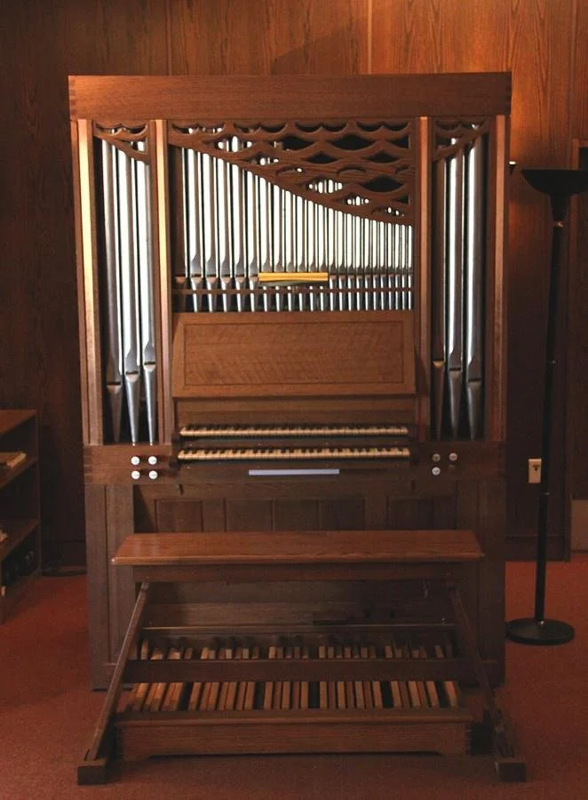 A pipe organ with wooden frame, metal pipes, and a keyboard in a room with wood-paneled walls and a standing lamp.