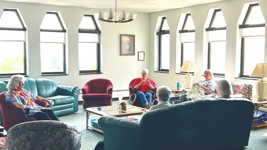 A group of elderly people sitting in a brightly lit room with multiple windows, some reading or relaxing, with furniture including sofas and armchairs.