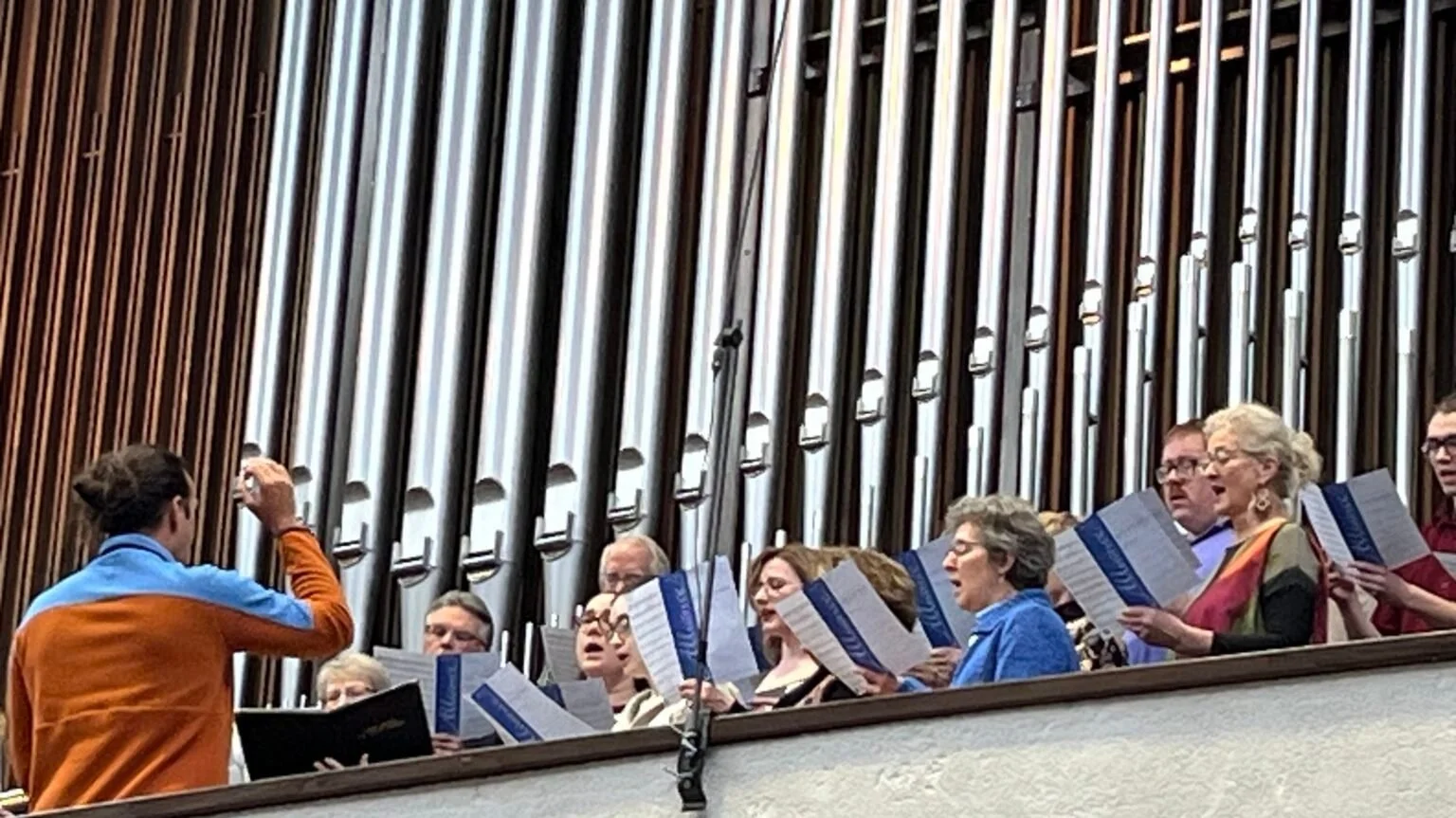 A choir singing in a concert hall with a pipe organ in the background.