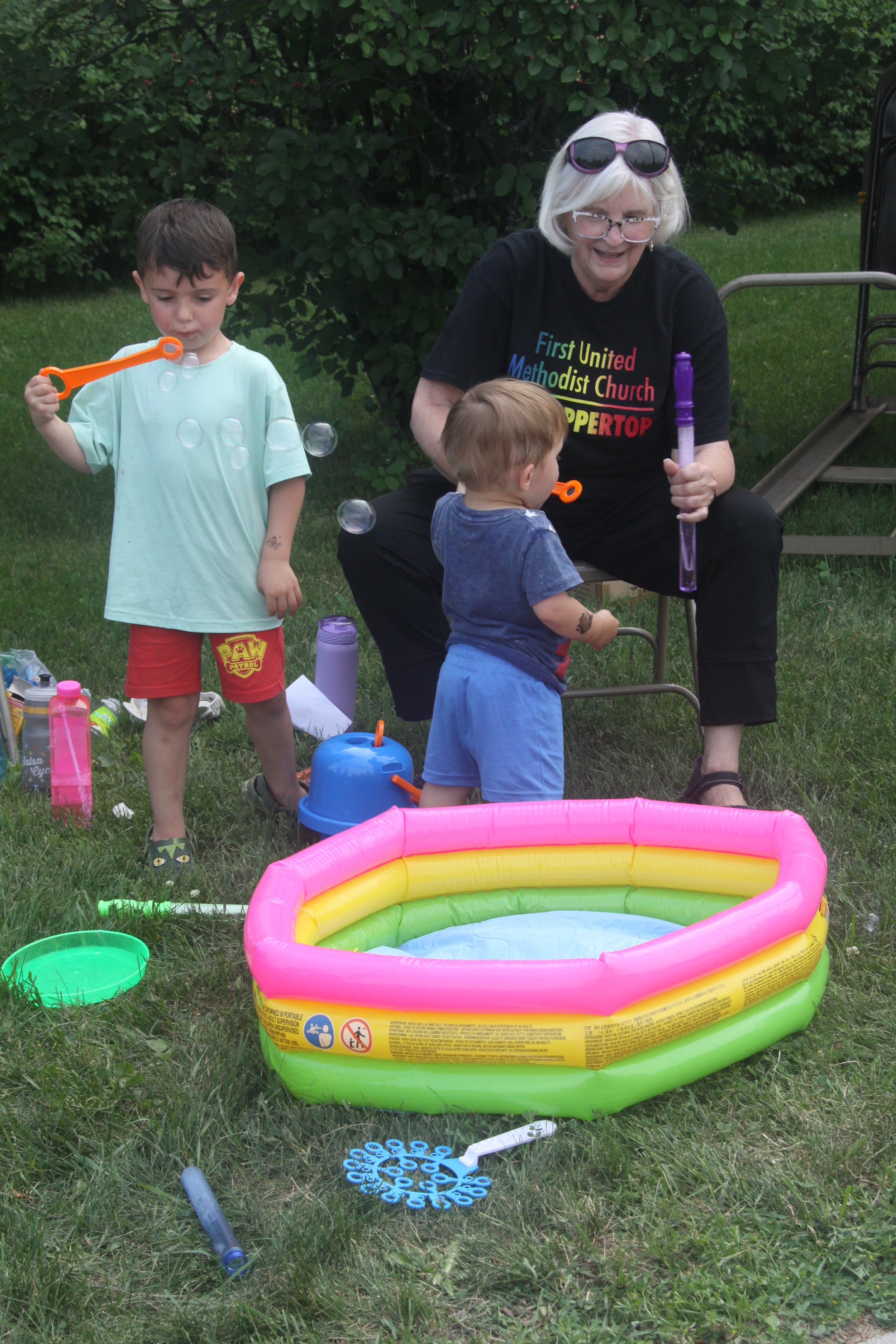 An older woman and two young boys enjoy a summer day outdoors with a small inflatable kiddie pool, bubbles, and water toys.