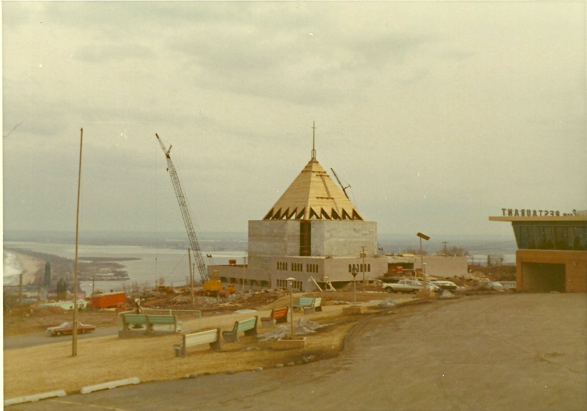 First United Methodist during construction with a conical roof, scaffolding, and cranes, located along a waterfront with a few parked cars and benches in the foreground.