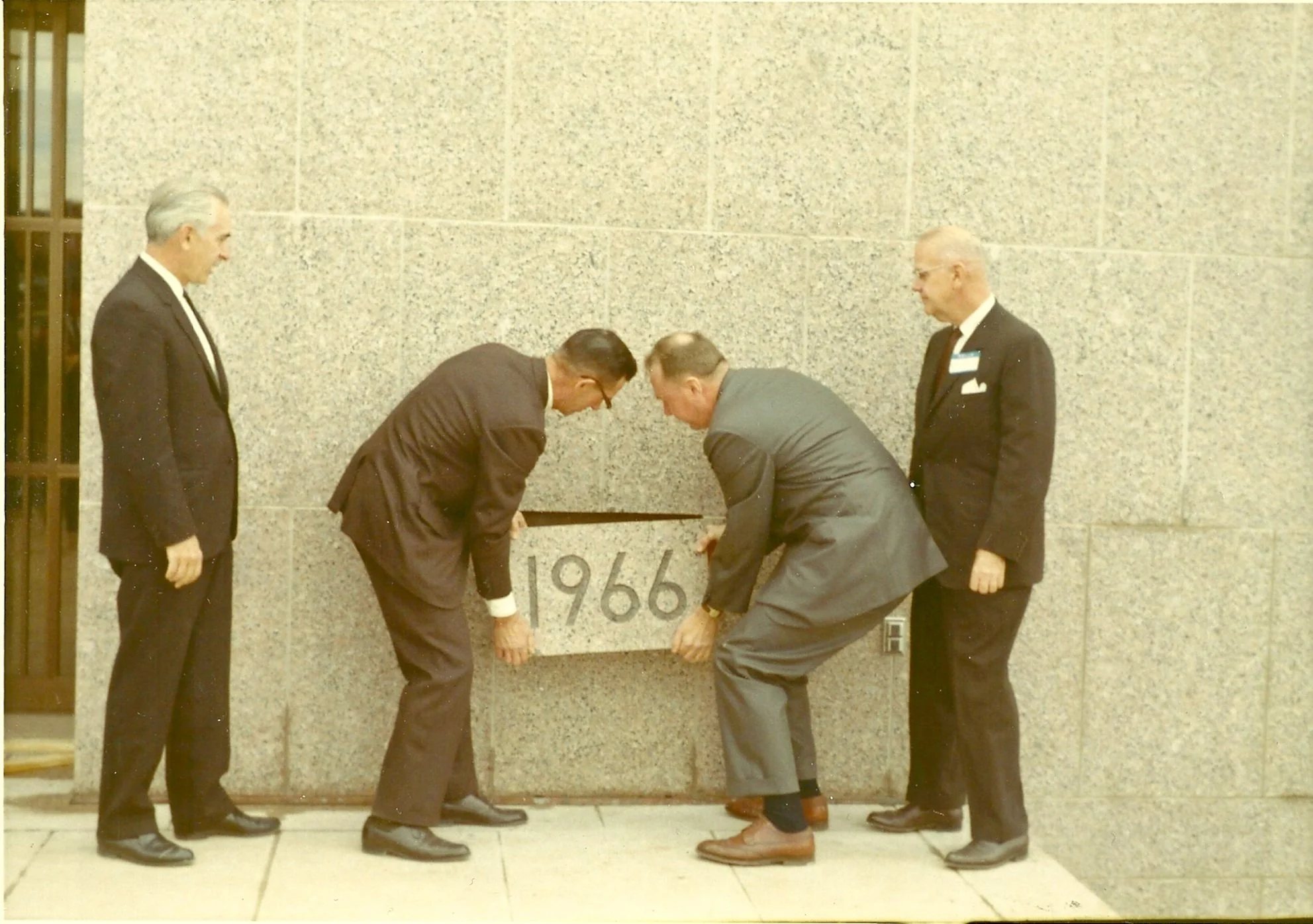 Four men in suits are unveiling a plaque with the year '1966' on it in front of a gray brick wall.
