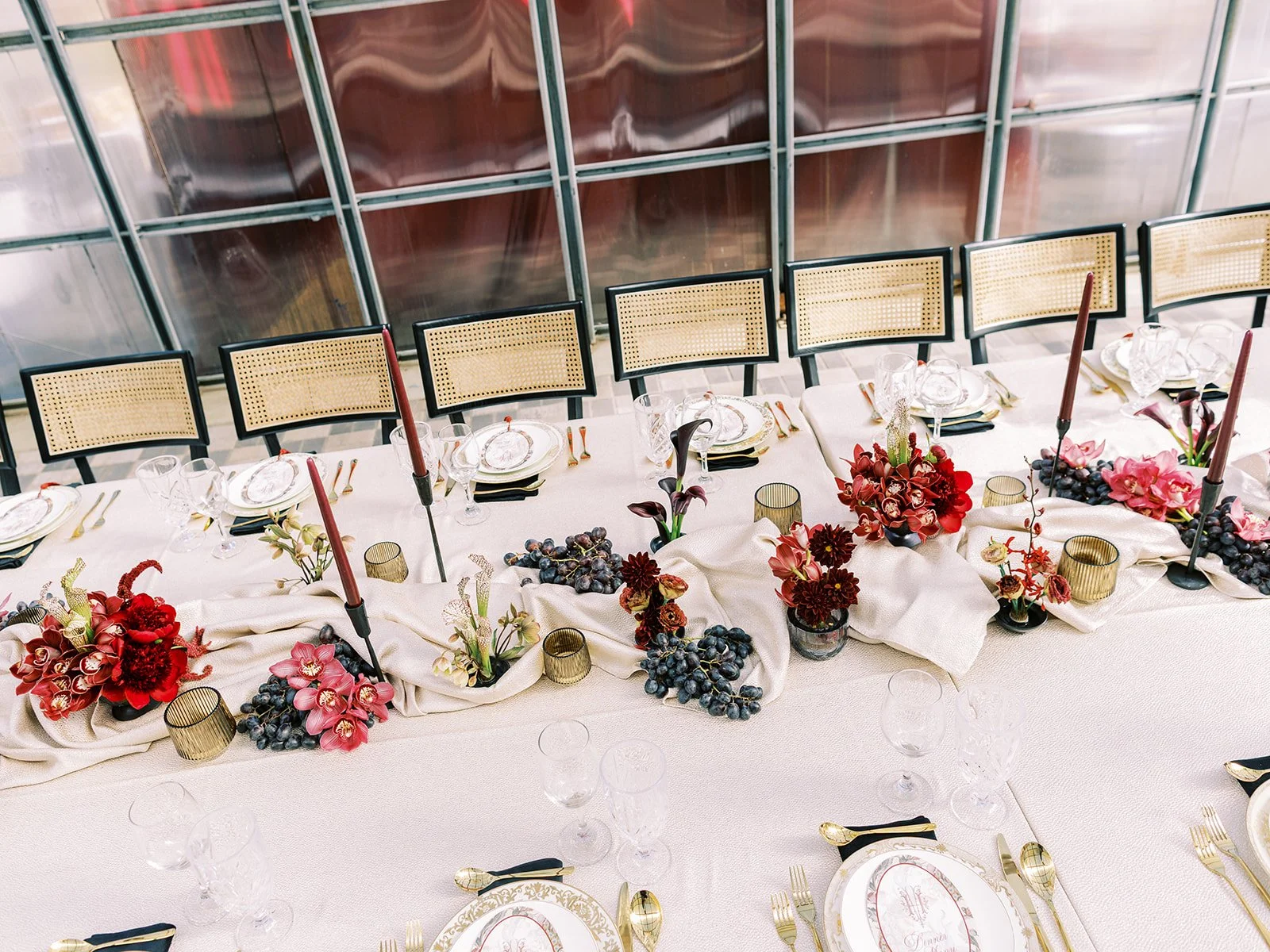 Elegant dining table with white tablecloth, decorated with red and pink flowers, grapes, gold and black cutlery, gold-rimmed plates, and gold votive candles, set with multiple wine glasses and tall red candles in black holders. Chairs are positioned along the table.