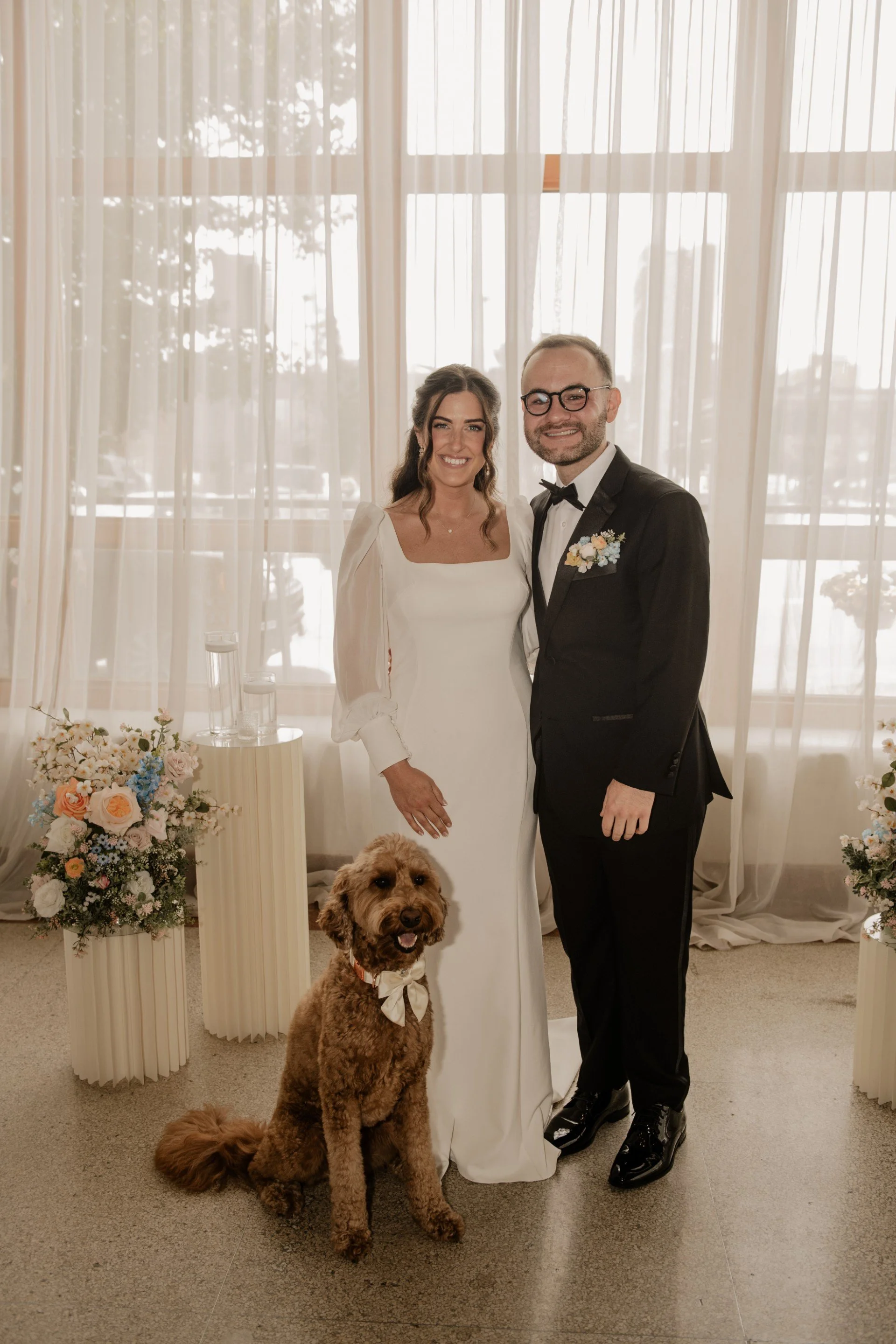 A bride and groom posing together in a wedding setting with a dog. The bride is wearing a white wedding dress with long sleeves, and the groom is dressed in a black tuxedo with a bow tie and glasses. They are standing in front of a backdrop of sheer curtains with natural light coming through. There are flower arrangements on either side, and a fluffy dog with a bowtie is sitting on the floor in front of them.