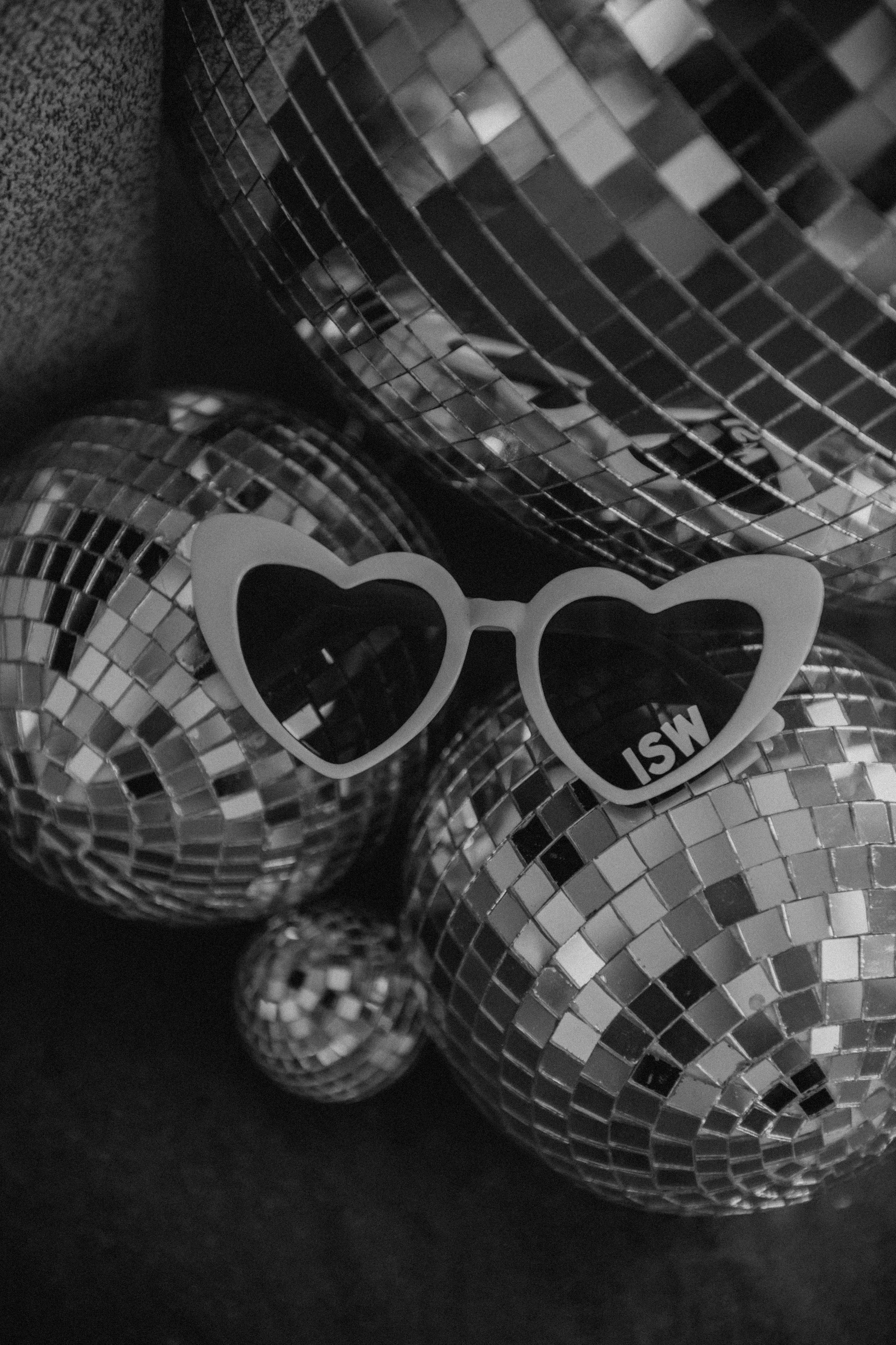 Moody black and white photo of disco balls with one pair of heart-shaped sunglasses resting on them. The disco balls have mirrored tiles, creating a reflective, shiny surface.