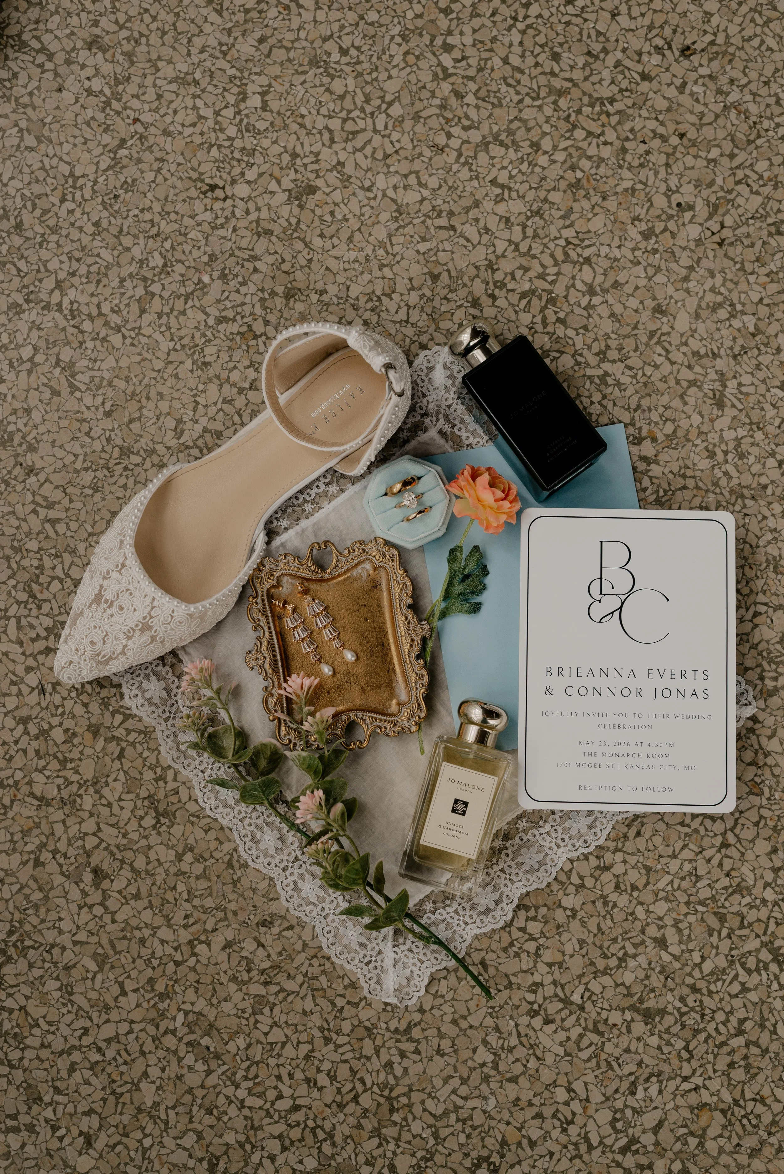 Wedding invitation, high heel shoe, jewelry, wine bottle, perfume, flower with green leaves, and decorative tray arranged on lace doily on a terrazzo floor.
