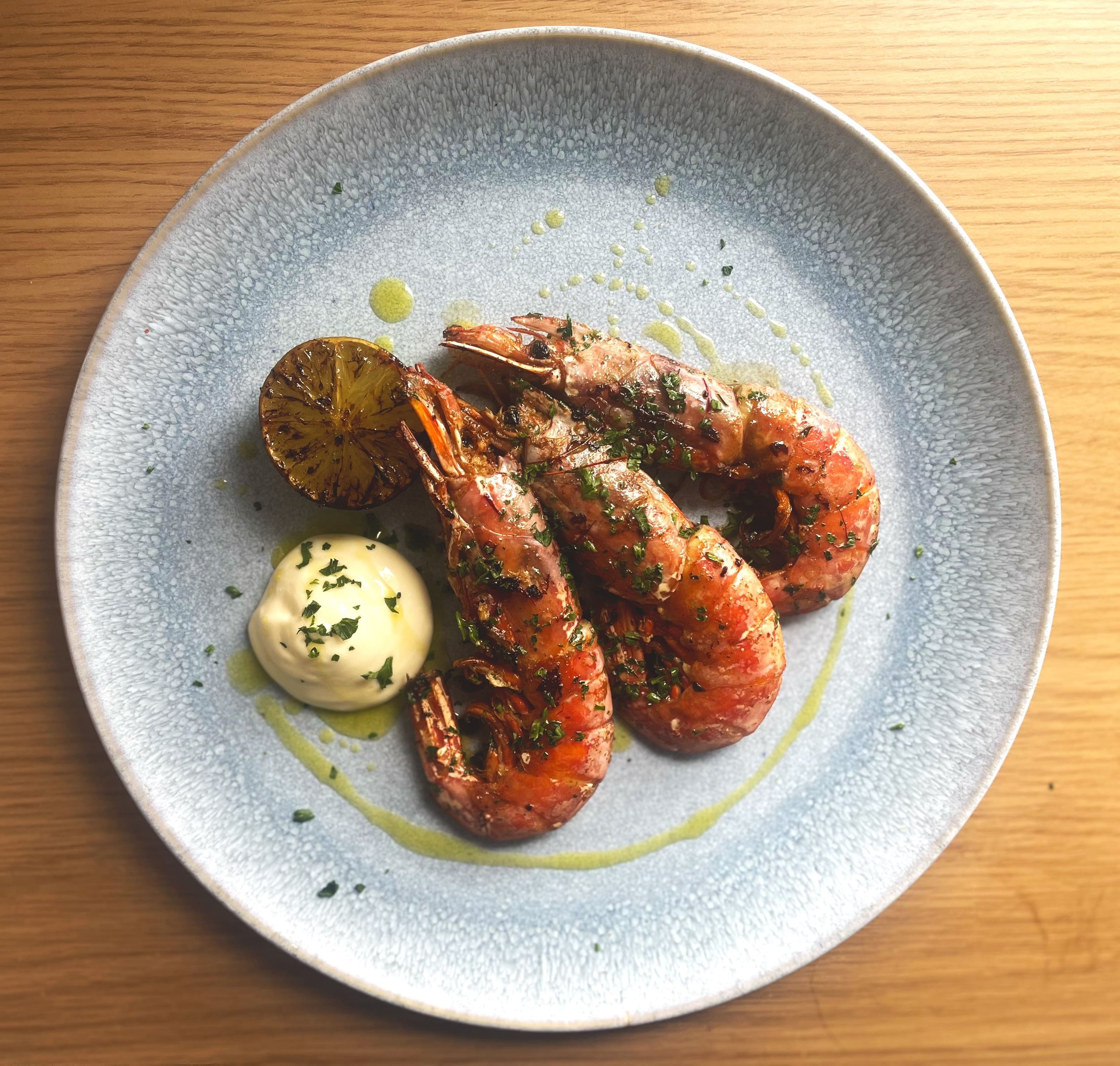 Plate of grilled shrimp garnished with chopped herbs, grilled lime half, and a dollop of creamy sauce on a speckled blue-gray plate.