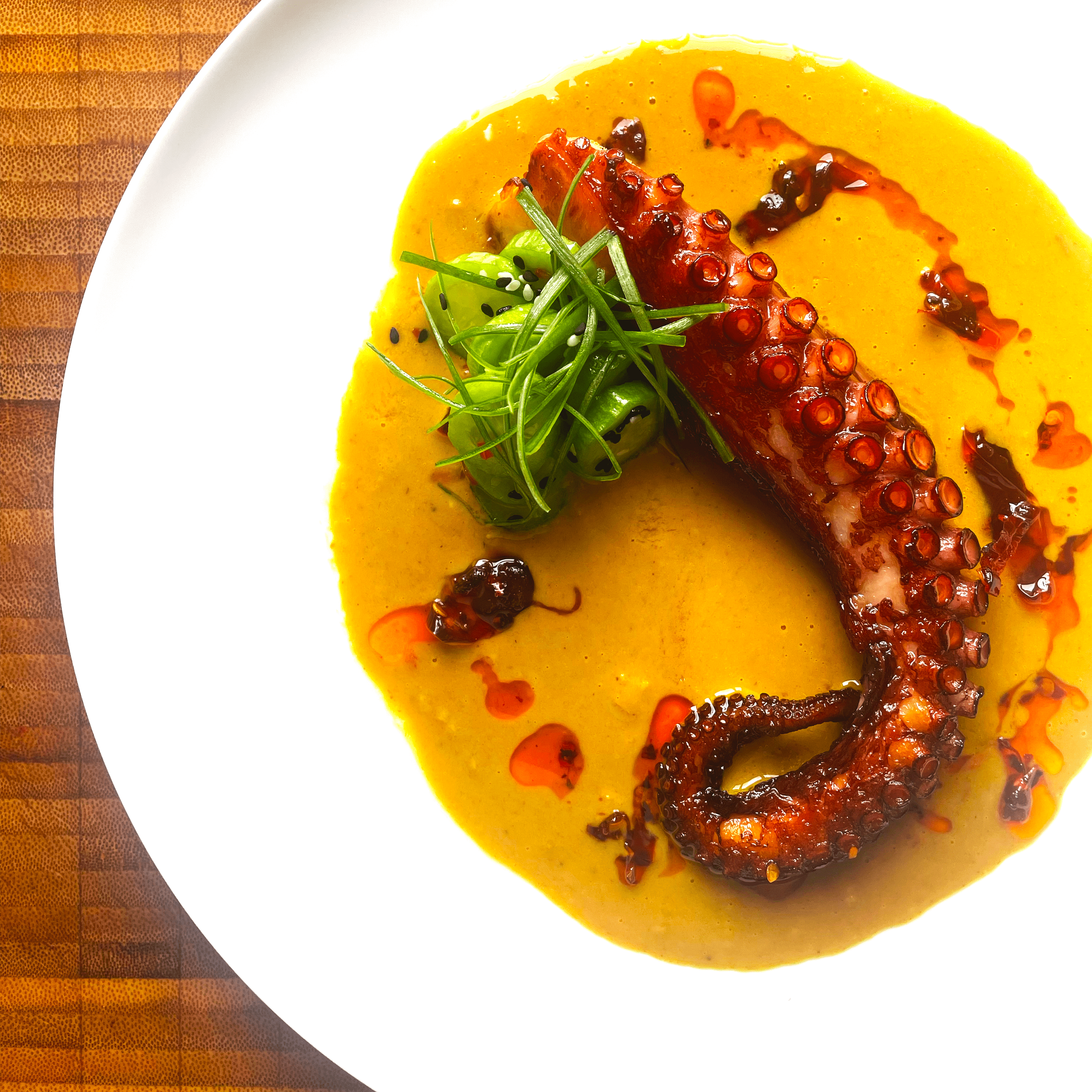 Cooked octopus tentacle served on a white plate with yellow sauce, garnished with green herbs, black sesame seeds, and drizzled with chili oil.