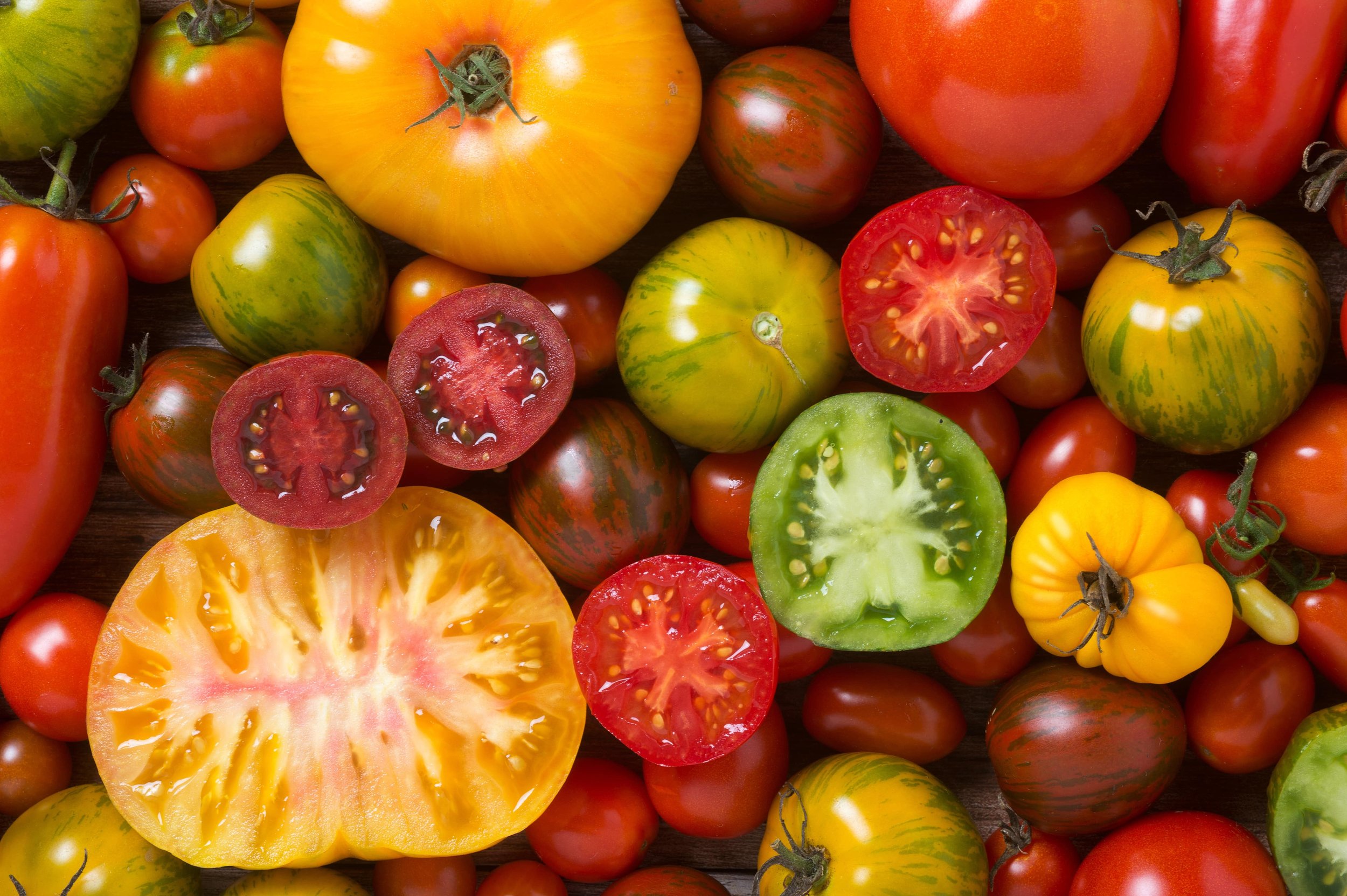 Assorted heirloom tomatoes in various colors, shapes, and sizes, some sliced open to reveal the interior, placed on a wooden surface.