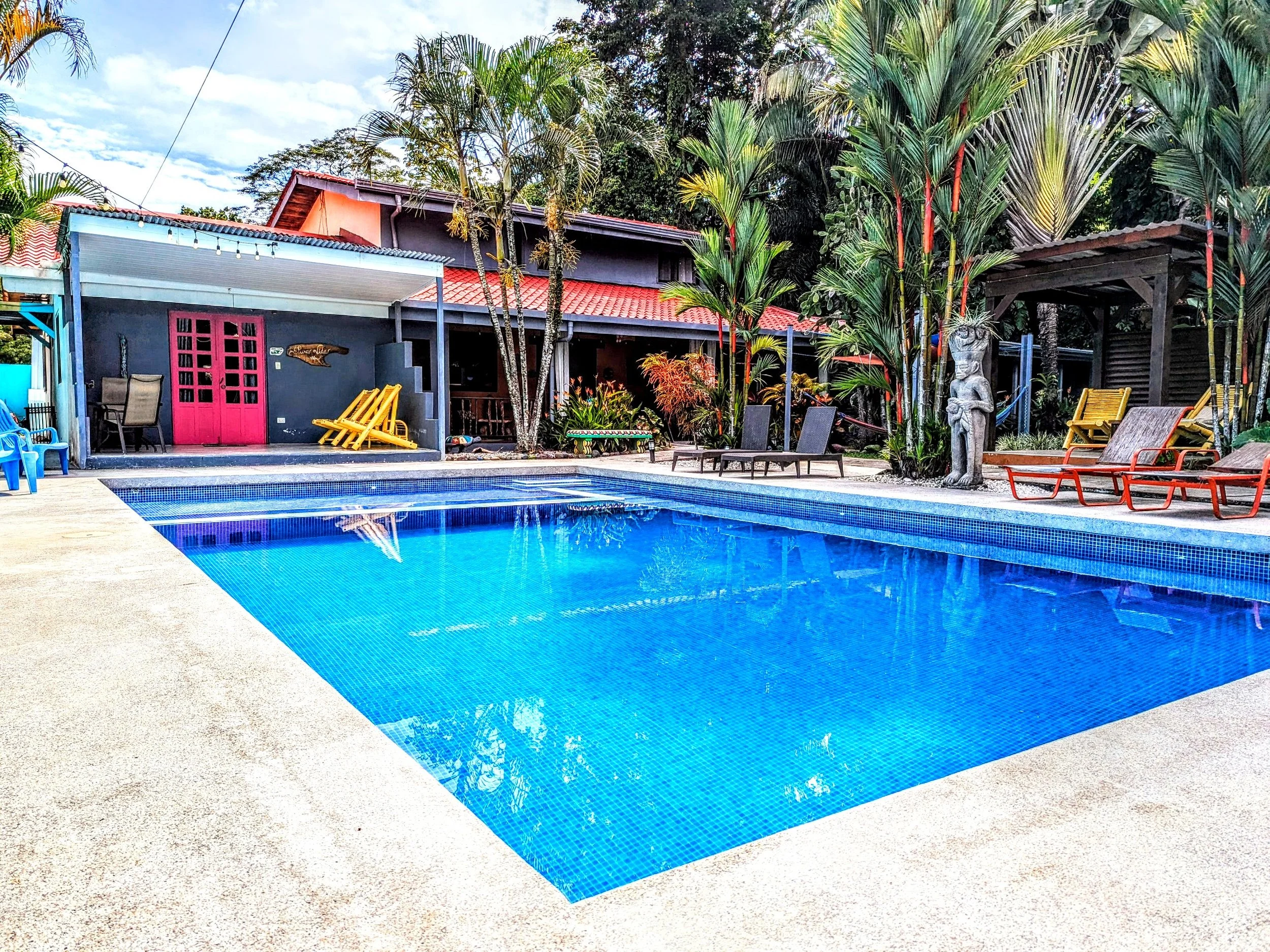 An outdoor swimming pool with blue water, surrounded by lounge chairs, tropical plants, and a house with pink double doors and a red tile roof in the background.