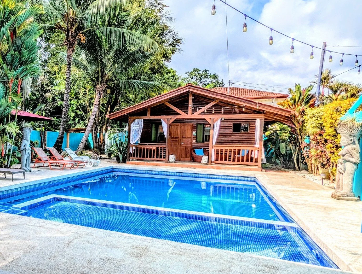 A backyard swimming pool surrounded by tropical plants and trees, with a wooden cabana in the background and string lights overhead.
