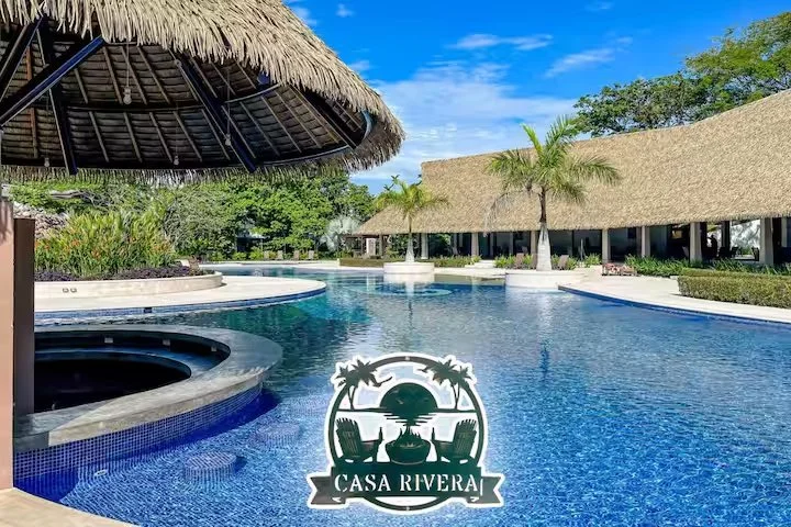 Luxury swimming pool area at Casa Rivera resort with thatched-roof cabanas, palm trees, and lush greenery, under a partly cloudy sky.