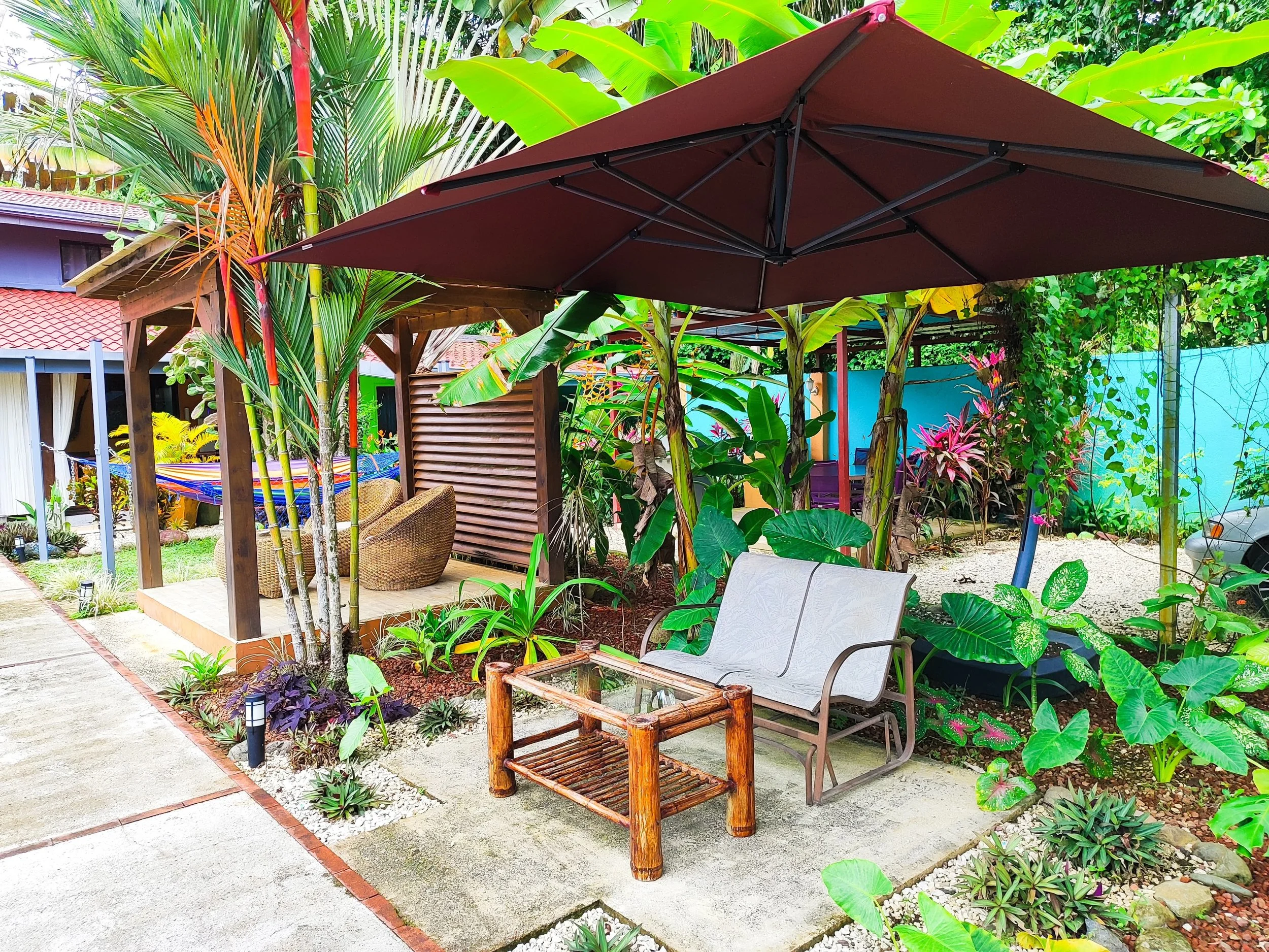 A cozy outdoor patio with a large brown umbrella, a wooden and glass coffee table, a white metal bench, surrounded by lush tropical plants, trees, and vibrant flowers, featuring a wooden shaded seating area with a hammock in the background.