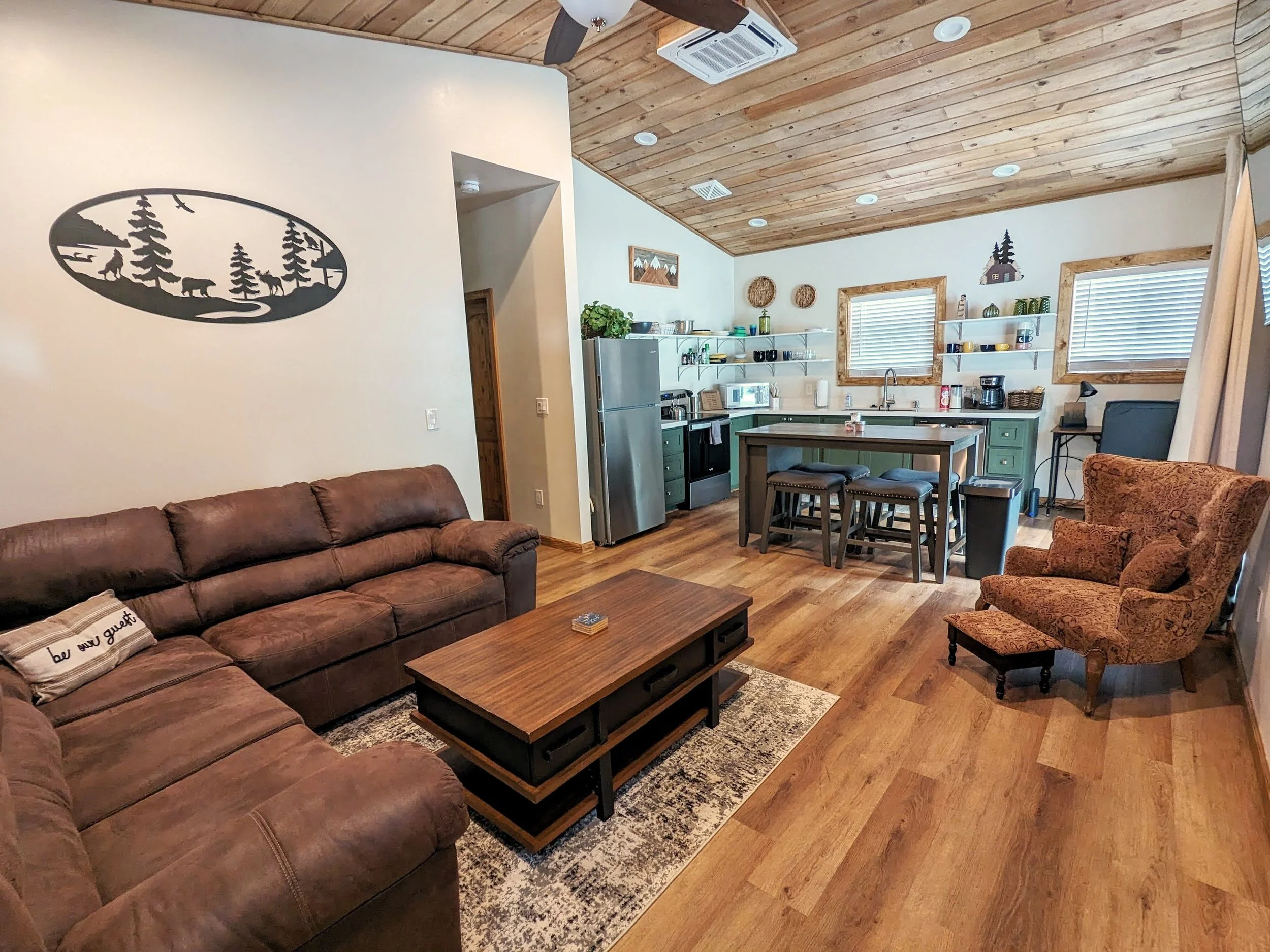 Living room with brown sectional sofa, wooden coffee table, patterned armchair with matching footstool, and an open kitchen in the background featuring green cabinets, open shelves, a stainless steel refrigerator, and a dining table with four stools.