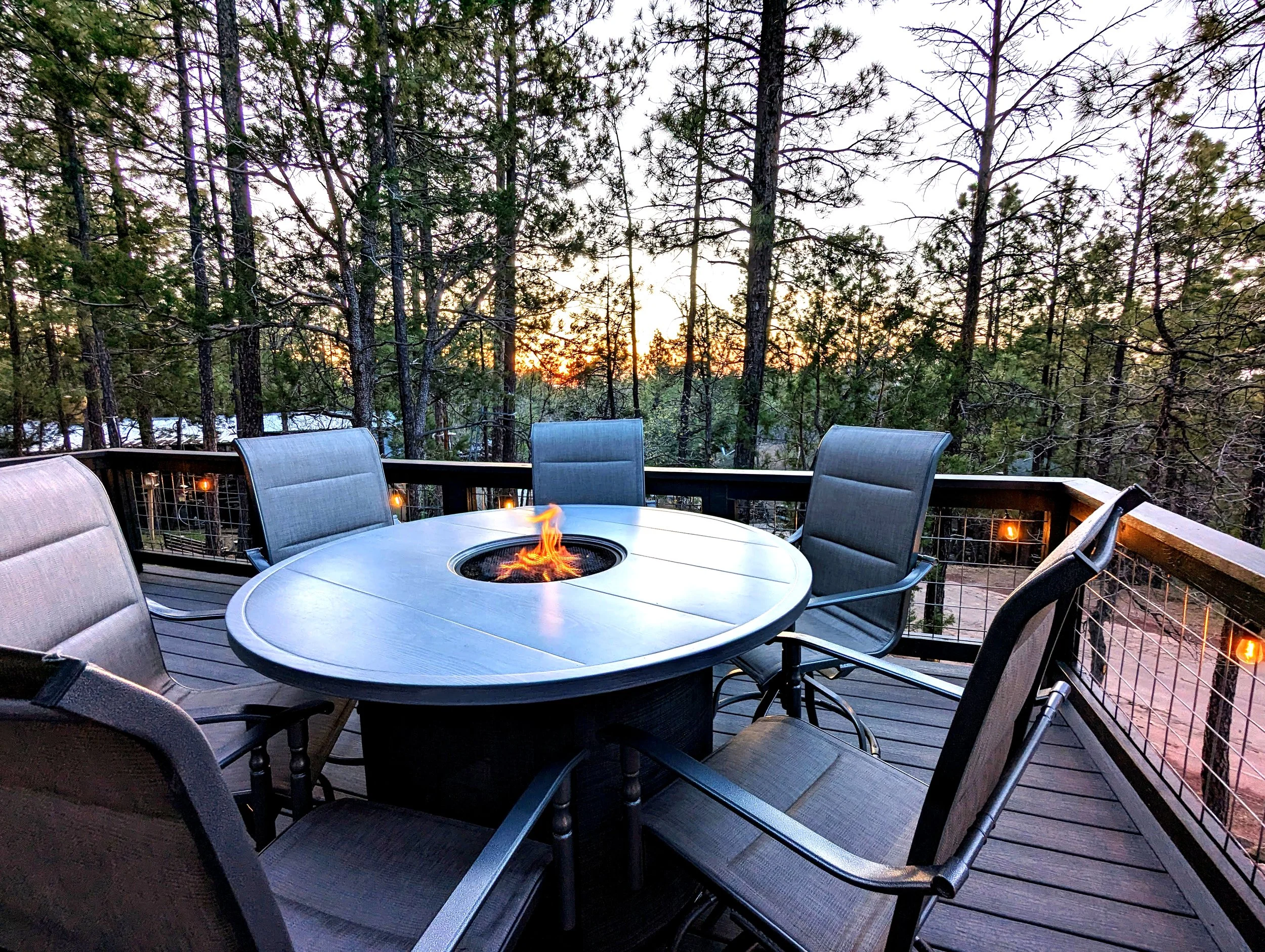 A round outdoor patio table with a built-in fire pit at the center, surrounded by six chairs, on a wooden deck with a forest and sunset in the background.